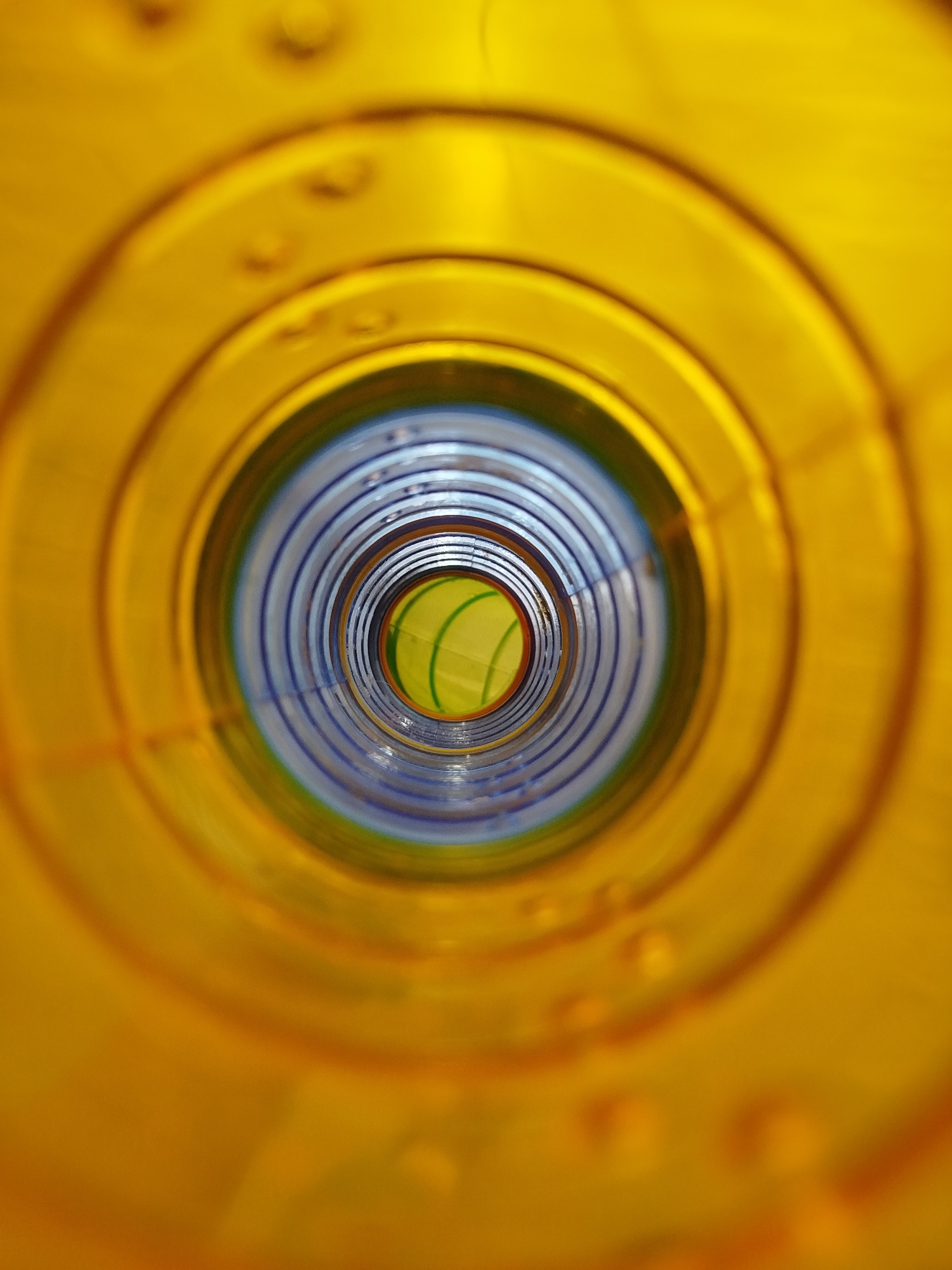 The image shows a close-up, abstract view looking down the interior of a yellow plastic tube or pipe. The tube has concentric rings and appears to be made of translucent yellow plastic. In the distance, the tube narrows, revealing inner layers of blue and green material. There are no people, actions, or interactions visible. The environment is entirely confined within the interior of this manufactured object, with no indication of its location in Kremenchuk, Ukraine, or any other specific place.