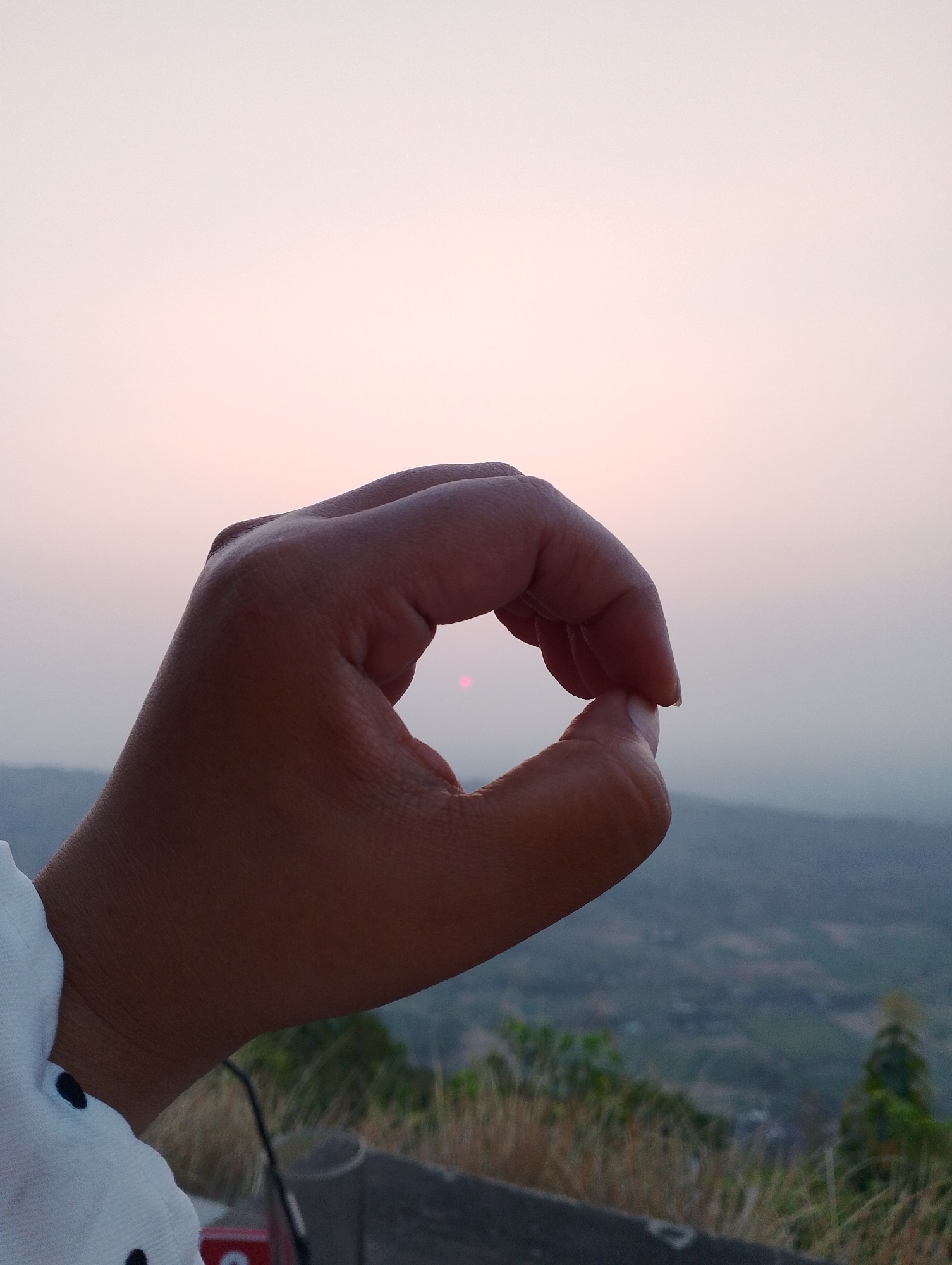 A person's hand is held up in the foreground, forming an "okay" gesture with their thumb and index finger. Through the circular opening created by the fingers, a small, reddish-orange sun is visible, partially obscured by haze. The hand is brown-skinned and appears to be wearing a white shirt with black polka dots on the sleeve, which is visible in the lower left corner. In the background, a hazy, mountainous landscape stretches out under a pale, overcast sky. The scene is likely a scenic overlook, possibly in Srimulyo, Indonesia, given the mountainous terrain and the presence of vegetation. The overall impression is one of a peaceful moment captured during either sunrise or sunset.