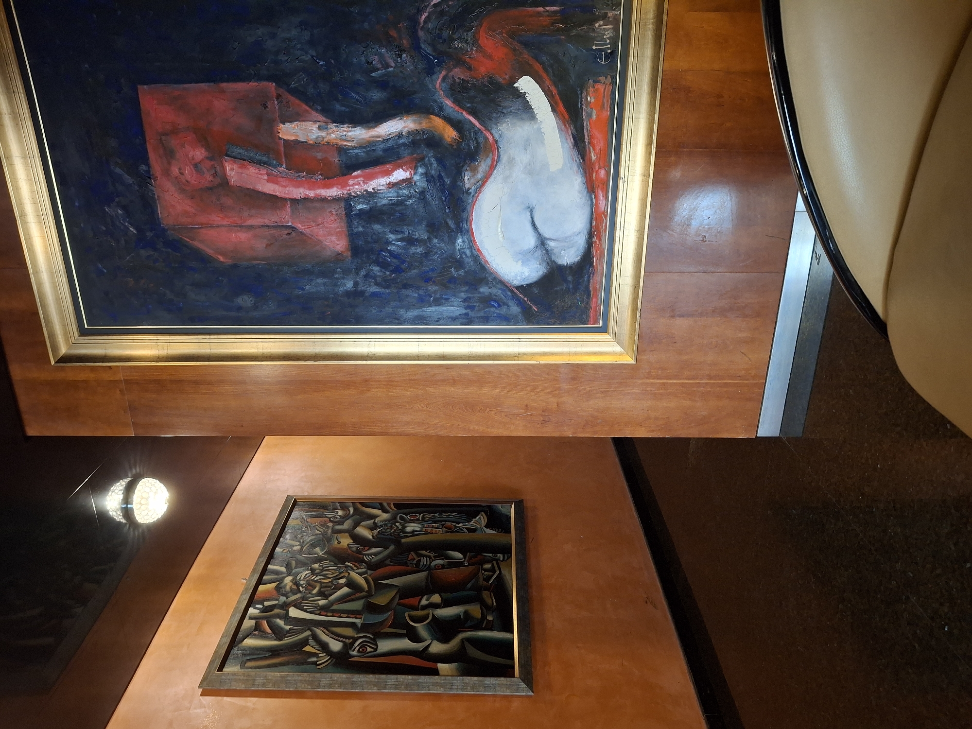 This media file captures an indoor setting displaying two distinct framed artworks, with the overall image appearing rotated clockwise.
In the upper section of the image, a large, rectangular oil painting is prominently displayed within a gleaming gold frame. The artwork features a dark, deep blue-black background with visible brushstrokes, indicating a rich texture. On the left side of the canvas, a dominant, angular red block or geometric form is rendered with thick impasto. To its right, a smooth, light grey human form, specifically depicting the lower torso and buttocks, is outlined in a vivid red. Dynamic, sweeping brushstrokes in red and off-white connect and extend from these central elements. A stylized signature, possibly "E.J. W." or similar, is faintly visible in the top right corner of the painting.
Below and to the right, a second, smaller painting is mounted on a textured, reddish-brown wall. This artwork, enclosed in a dark, simple frame, exhibits a dense, cubist or expressionist style. It portrays multiple stylized human figures or fragmented body parts, interwoven into a complex composition. The figures are rendered in earthy tones of browns, greys, reds, and blacks, characterized by sharp angles and prominent facial features.
The surroundings include polished wooden paneling, which serves as a backdrop for the upper painting and forms part of the wall structure. A round, illuminated light fixture with a honeycomb pattern is visible on a darker, reflective surface, possibly a ceiling or upper wall, casting a soft glow and reflections. To the far right, a curved section of a light-colored, possibly beige leather chair or sofa is partially visible. The floor or a lower wall section appears dark and glossy. The scene evokes a sense of a curated interior, such as an art gallery, a hotel lobby, or a well-appointed private space.