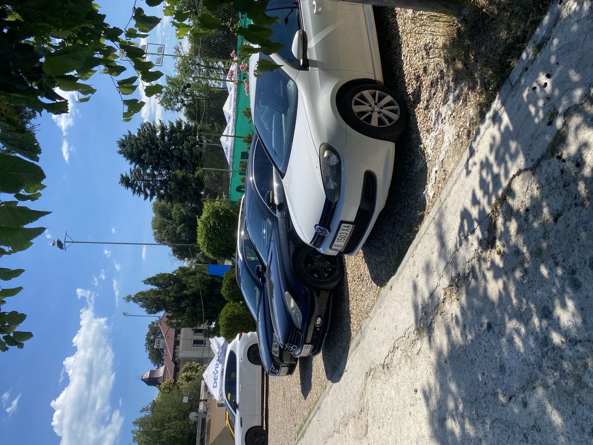 The image shows a parking lot with several cars. In the foreground, there is a white Volkswagen Golf. Behind it is a black Mercedes-Benz CLS. Further back, a black Volvo XC60 is parked. On the left, a white SUV is visible. In the background, there are trees, a building with a red roof, and tents with the "DEVIN" logo. The sky is blue with scattered clouds. This scene appears to be located at the Varna Sports Palace, a sports and entertainment complex in Varna, Bulgaria.
