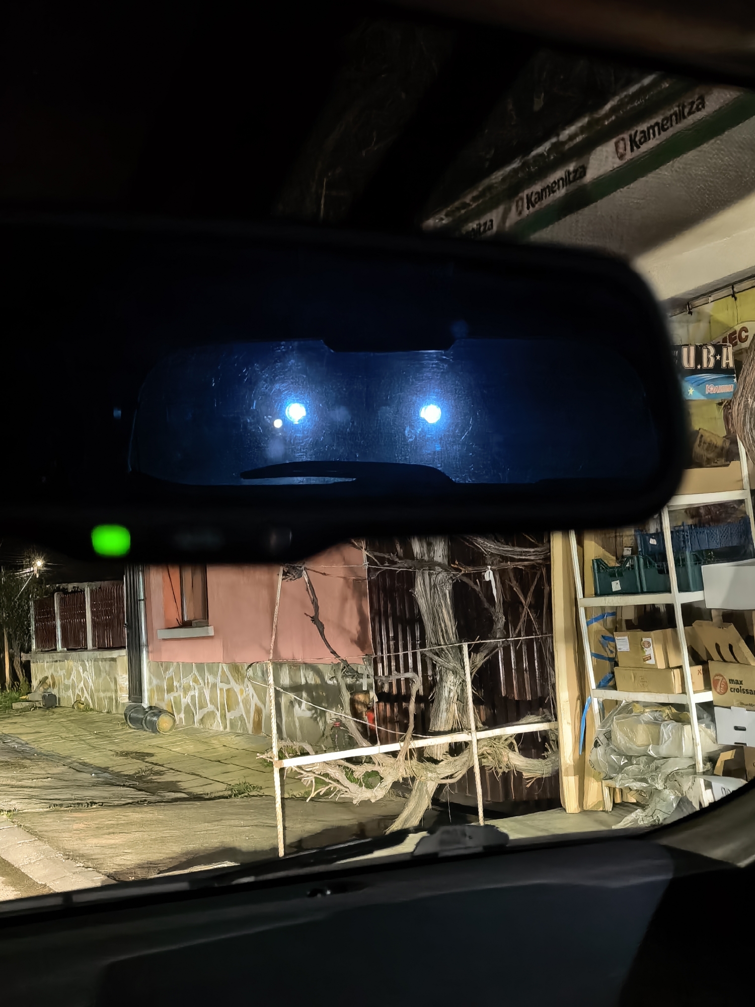 The image shows a view from inside a car through the windshield. The car's rearview mirror is visible, reflecting the headlights of an approaching vehicle. Outside the car, there's a building with a pink exterior and a stone base. A metal fence and some bare branches are in front of the building. To the right, there's a shop or storage area with shelving containing various boxes and containers. The location is Unknown, Unknown. The time is night. The scene is lit by headlights and artificial light sources.