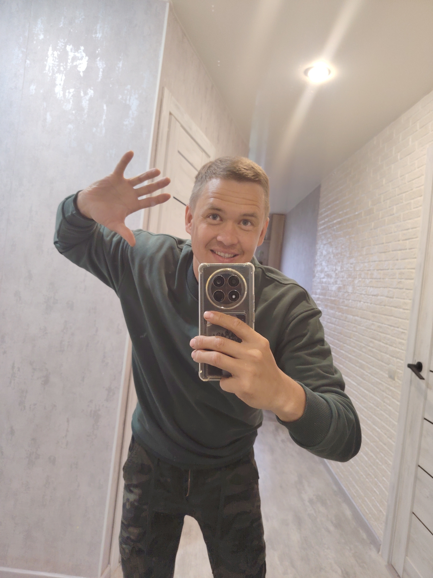 A mirror selfie shows a man standing indoors, likely in a hallway or room. He appears to be in his 30s or 40s, with short, light-colored hair and a friendly, broad smile that reveals his teeth. He is wearing a dark green crew-neck sweatshirt and dark-colored trousers with a subtle patterned design, possibly camouflage.
The man holds a smartphone with a clear, textured protective case in his right hand to capture the reflection. The phone's back features a prominent circular camera module with multiple lenses. His left hand is raised with the palm open, gesturing as if waving or saying hello.
The background, seen through the mirror, consists of an interior space. The wall behind him on the left features light grey wallpaper with a subtle, textured, possibly metallic pattern. On the right, the wall has an off-white, painted brick-like texture. A light-colored door with a dark handle is partially visible on the right. The ceiling is smooth and light, illuminated by a bright, recessed circular light fixture. The flooring is light-toned. The overall impression is a cheerful moment in a well-lit, modern indoor setting. No discernible text is visible on any objects or surfaces within the image. The provided context indicates the location is Chaikovsky, Russia.