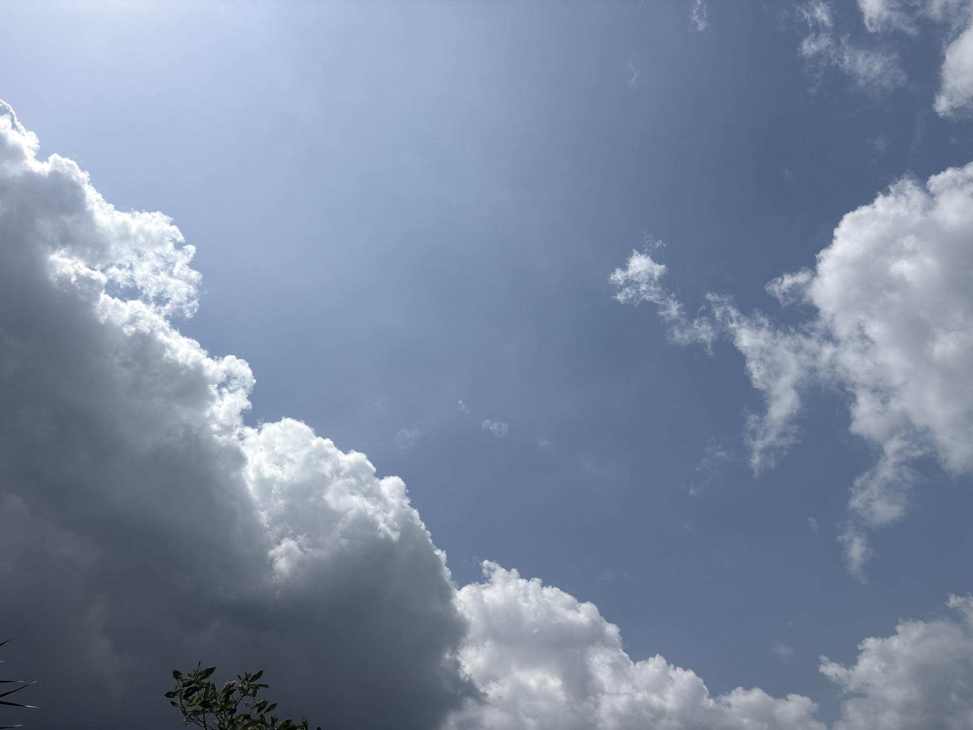 The image depicts a view of the sky in Ikot Ekpene, Nigeria. The sky is predominantly light blue, with the sun shining through a hazy atmosphere. Large cumulonimbus clouds are present on the left and right sides of the frame. The clouds on the left appear darker and denser than those on the right. A few sparse clouds are also visible in the center of the image. Some green foliage is visible at the bottom of the frame, suggesting the presence of trees or vegetation below the camera's viewpoint. No people or man-made objects are evident in the image.