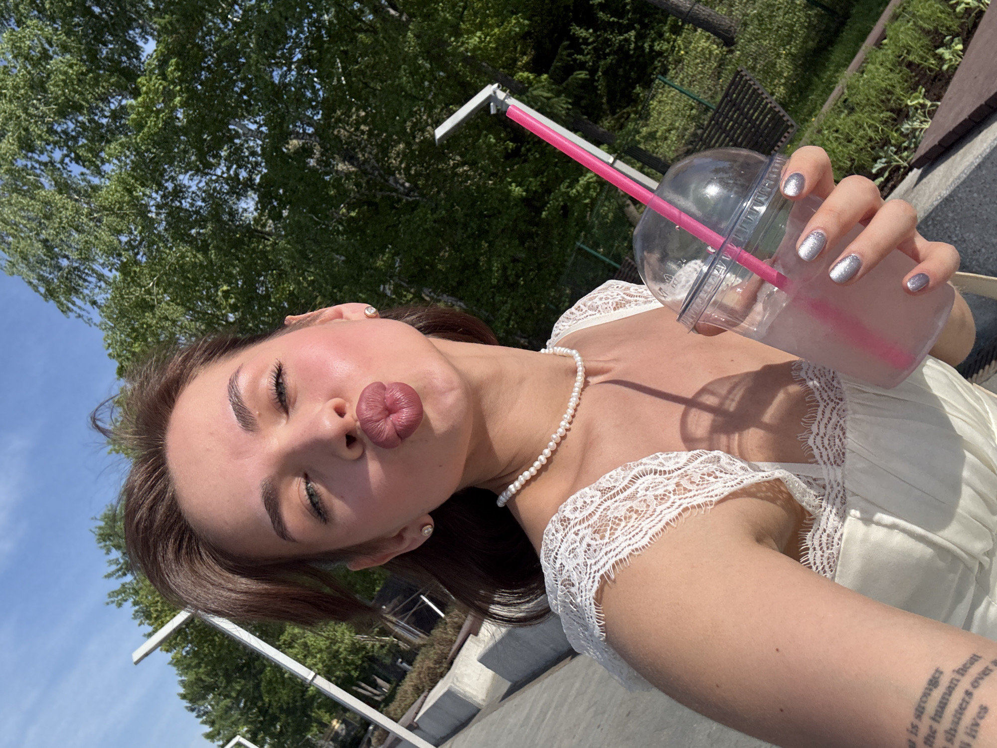 A young woman with brown hair, wearing a white dress with lace trim and a pearl necklace, is taking a selfie. She is puckering her lips towards the camera. She is holding a clear plastic cup with a pink liquid and a pink straw. She has silver glitter nail polish. A tattoo is visible on her arm. In the background are trees, blue sky, and what appears to be part of a modern outdoor structure. The location appears to be in the city of Perm, Russia, based on the architectural style and vegetation.