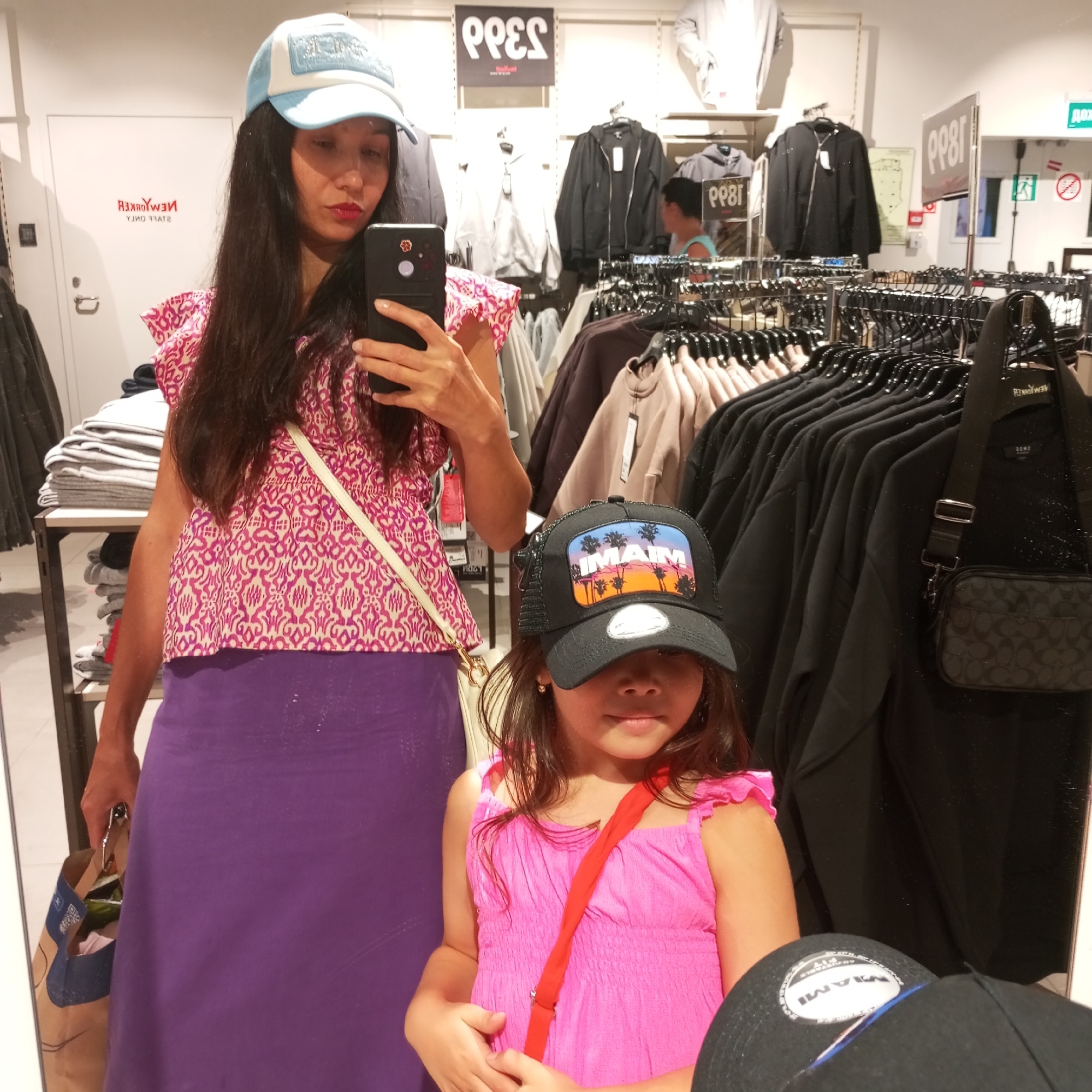 Two females are reflected in a mirror within a retail clothing store. The female in the foreground, on the left, is wearing a light blue baseball cap, a pink patterned top with puffy sleeves, and a long purple skirt. She is holding a black smartphone in her right hand, taking a selfie. A beige crossbody strap is visible over her left shoulder. She is also carrying a brown paper shopping bag in her left hand. To the right of the first female, a younger female is visible, wearing a black trucker hat with a tropical scene graphic, and a pink sleeveless dress with ruffled straps. She is looking towards the mirror. Various racks of clothing are visible in the background, with hangers and garments. Signs with text are also present, including one that reads "PP8T". A black bag with a patterned strap is hanging from a clothing rack on the right side of the image. The overall environment suggests a commercial setting, likely a clothing boutique or department store. The presence of specific retail signage and clothing displays indicates an indoor shopping environment. No explicit indication of the city of Sochi, Russia, is present in the image itself.