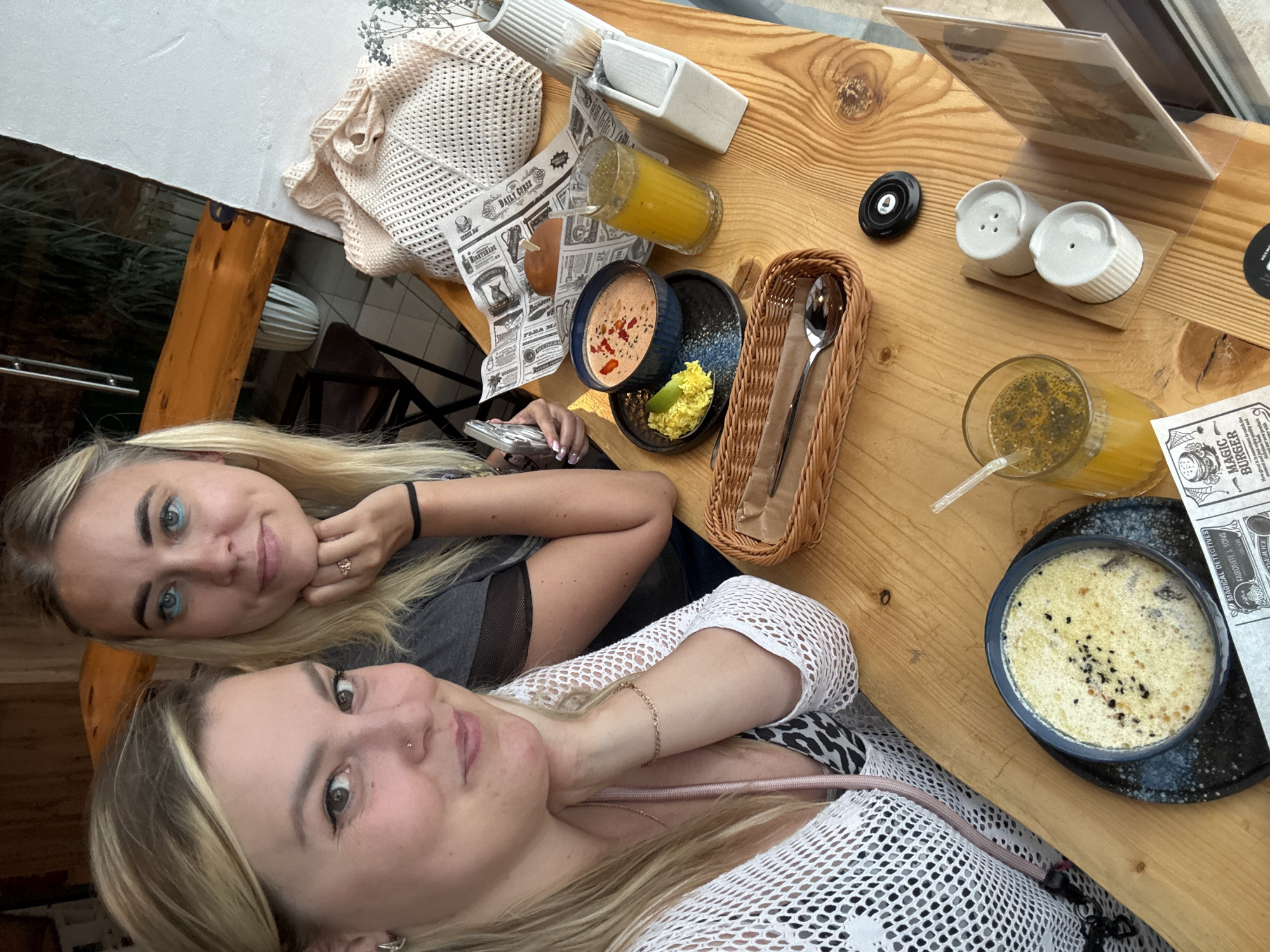 Two young Caucasian women are seated at a wooden table, posing for a selfie in what appears to be an outdoor cafe or restaurant. The woman in the foreground, on the left, has blonde hair and is wearing a white mesh top over a patterned undergarment. She is resting her chin on her hand, looking towards the camera with a slight smile. The woman in the background, on the right, also has blonde hair and is wearing a dark grey t-shirt. She is holding a silver smartphone in her right hand, with her left hand resting on her chin.
On the table are several food and drink items. To the left, there is a bowl of creamy soup with black sesame seeds sprinkled on top. Next to it is a glass of yellow juice with a straw, and a salt and pepper shaker set on a small wooden base. In the center, there's another bowl containing a reddish soup with visible chunks, accompanied by a small portion of yellow rice and a wedge of lime. Beside this, another glass of yellow juice with a straw is visible. A woven basket containing a spoon is also on the table, along with a bun and a folded newspaper. A cream-colored knitted bag is placed in the background, to the right.
The background of the image shows part of the cafe's interior, with a wooden beam and a white wall. Through a glass panel, some greenery and outdoor seating can be observed. The overall lighting suggests it is daytime. The presence of a newspaper with what appears to be Russian text indicates the location is likely in Russia, and specifically the city of Chaikovsky, given the prompt.