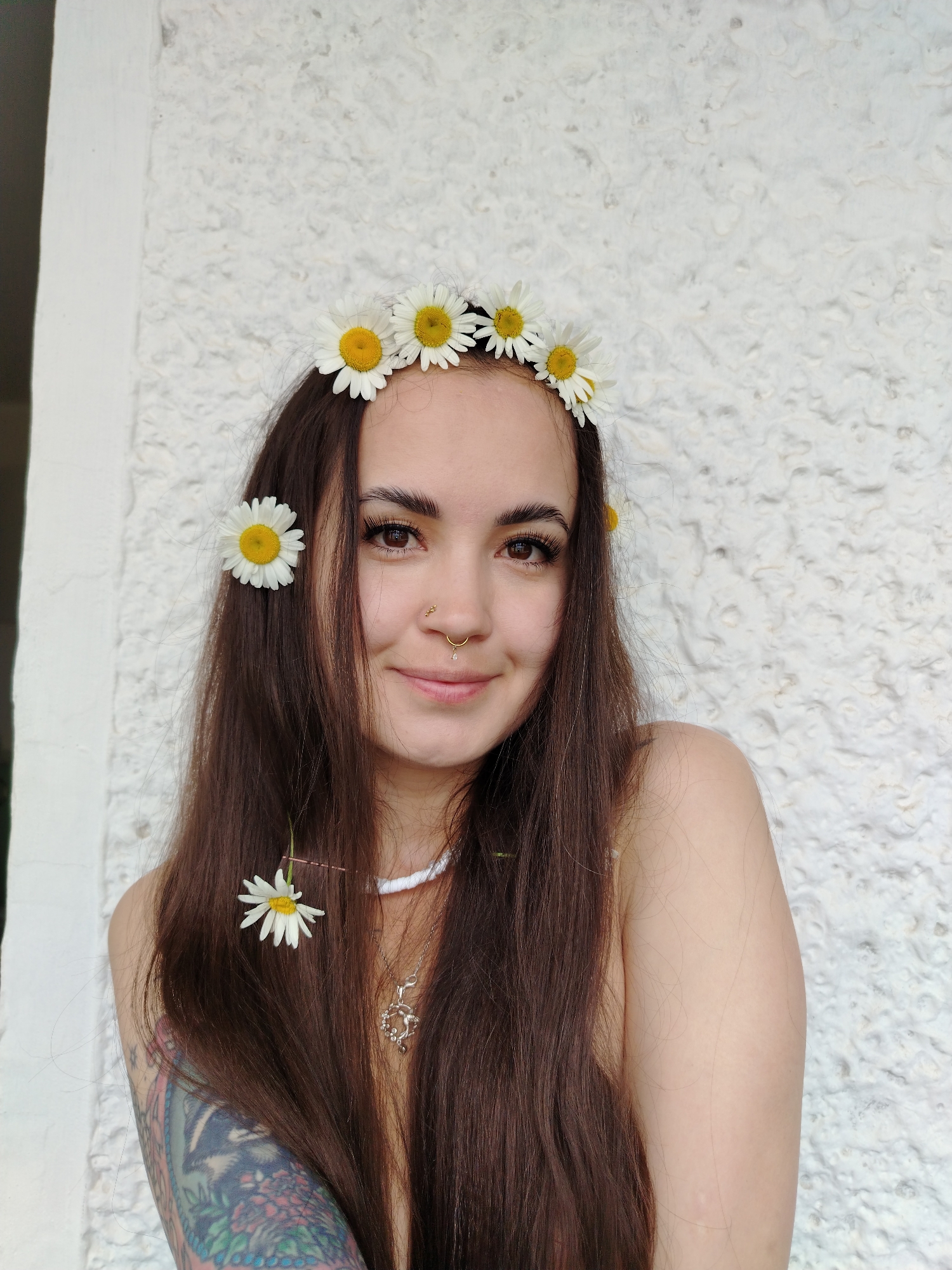 A young woman with long, dark hair adorned with a daisy crown and a single daisy tucked behind her ear smiles at the camera. She has a nose ring and a septum piercing. She is wearing a white beaded necklace with a silver pendant. A tattooed arm is visible on her left side. The background is a textured white wall. The image is a portrait taken outdoors, possibly in the city of Noviy, Russia.