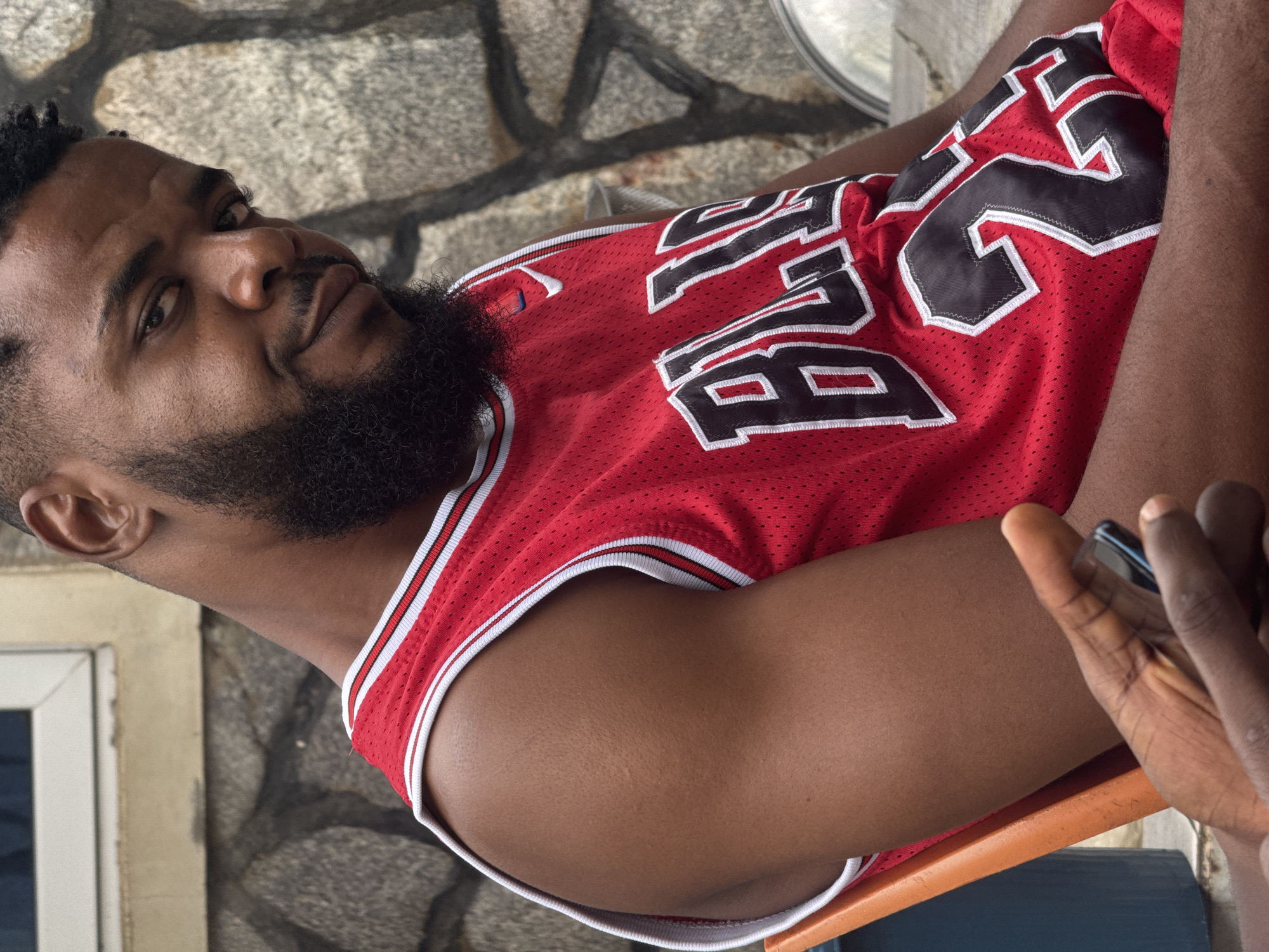 The image shows a close-up, outdoor portrait of a man wearing a red Chicago Bulls basketball jersey with the number 23 on it. He has dark skin, a beard, and is looking at the camera with a slight smile. The man is seated on an orange chair, and behind him is a stone wall and a window with a white frame. Another person's hand holding a smartphone is visible in the lower left corner of the frame. The setting appears to be a residential area, possibly in Akwanga, Nigeria, given the architecture and natural light. The overall impression is casual and relaxed.