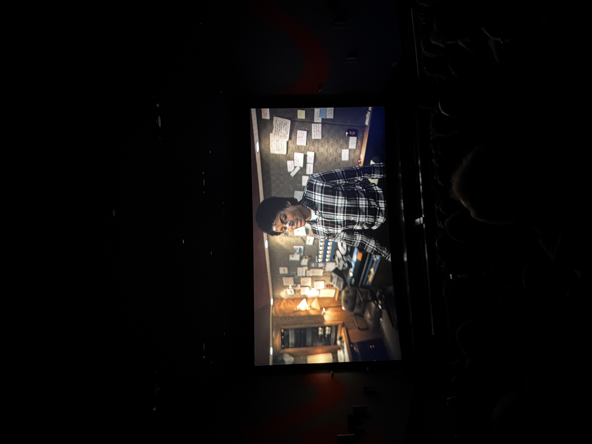 A dark environment, likely a cinema or darkened room, frames a brightly illuminated screen. The screen is rotated ninety degrees clockwise, displaying an indoor scene from what appears to be a film or television show.
On the screen, the central subject is a person, likely male, with dark skin and short dark hair, wearing a plaid shirt. They are looking directly towards the viewer with a slight, friendly smile. Behind the person, a wall is covered extensively with numerous sticky notes or pieces of paper, suggesting an office or studio setting, perhaps for planning or investigation. In the foreground of the screen's image, a desk or console with various equipment, possibly related to audio or recording, is visible along with a small lamp emitting a warm glow. The interior lighting within the screen's scene is warm and relatively dim.
The surrounding area outside the screen is almost completely black, though faint outlines on the right side suggest the heads or shoulders of audience members or other objects. There is a subtle, curved reddish glow or reflection visible both above and below the displayed screen. No legible text is discernible from the sticky notes or any other part of the image. The overall activity depicted is the viewing of media in a darkened venue.