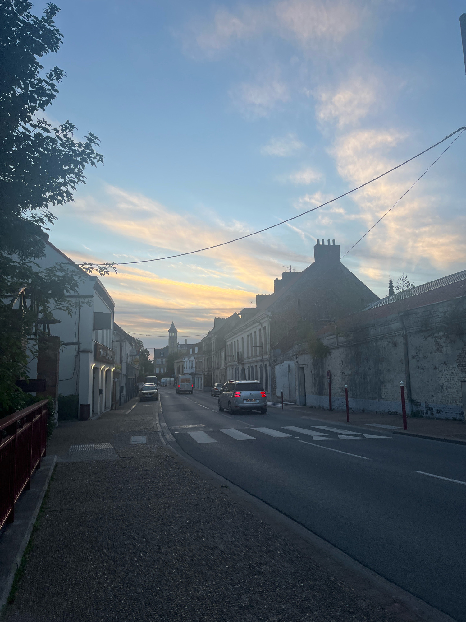 A street scene in Arques, France, at dusk. The image looks down a street lined with buildings on both sides, with cars driving on the road. The sky is a mix of blue and pastel-colored clouds, indicating either sunrise or sunset. A tree on the left side of the image partially obscures the view of the buildings. The buildings appear to be a mix of residential and commercial properties, some with lights on. A church tower is visible in the distance. A crosswalk is painted on the road, and several cars are driving on it. The overall mood of the image is calm and serene.