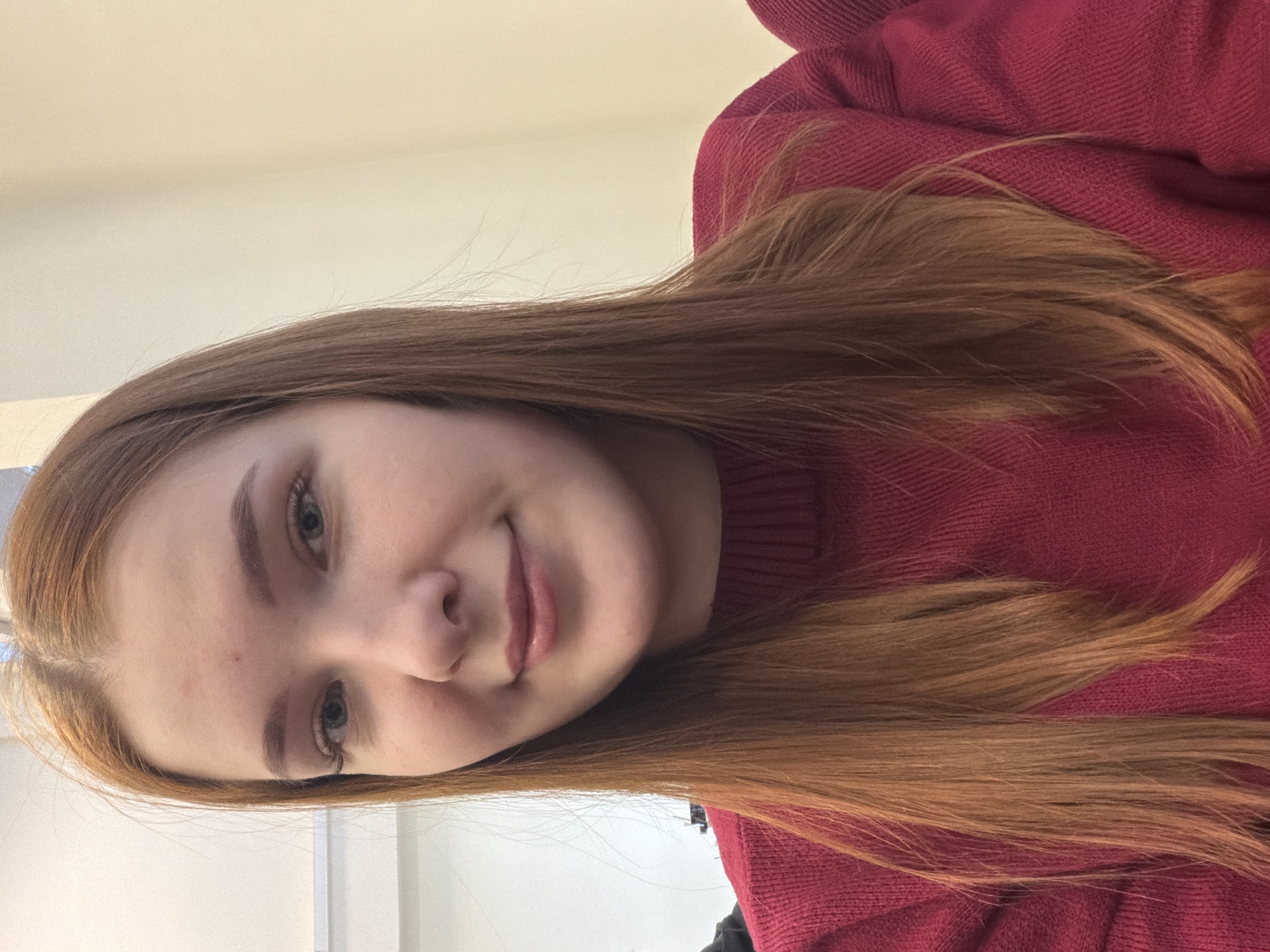 A portrait-oriented image, rotated 90 degrees clockwise, features a young woman indoors. She has long, straight, reddish-brown hair that cascades over her shoulder and rests against a red, possibly knitted, sweater. The woman looks directly at the camera with a slight, gentle smile, conveying a calm and friendly expression. Her eyes are light-colored, and she has fair skin. The background is a plain, light-colored wall or ceiling, indicating an indoor setting. There are no specific cues to the time of day or weather within the image itself. No text is visible. The provided location context is Chaikovsky, Russia.