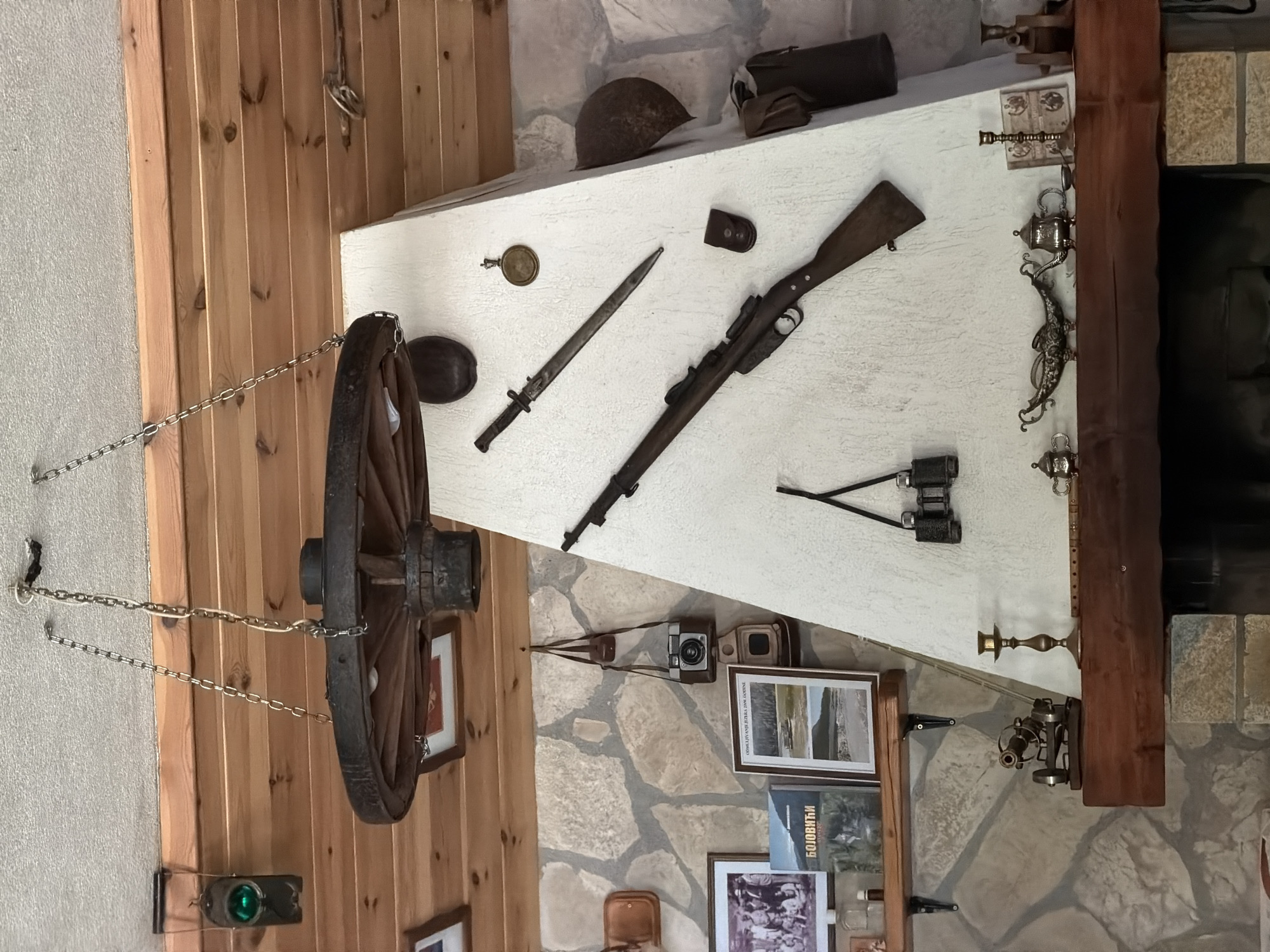 The image depicts an interior space in Njegovuđa, Montenegro, with a mantelpiece displaying various historical and decorative items. A wagon wheel hangs on the left, suspended by chains. The mantelpiece itself holds a collection of objects, including a military helmet, a rifle, a bayonet, binoculars, and what appears to be military pouches. Below the mantelpiece, there are framed photographs and what looks like a vintage camera. The walls are a mix of wood paneling and stone, adding to the rustic aesthetic. A fireplace is visible on the right, framed by a wooden beam.