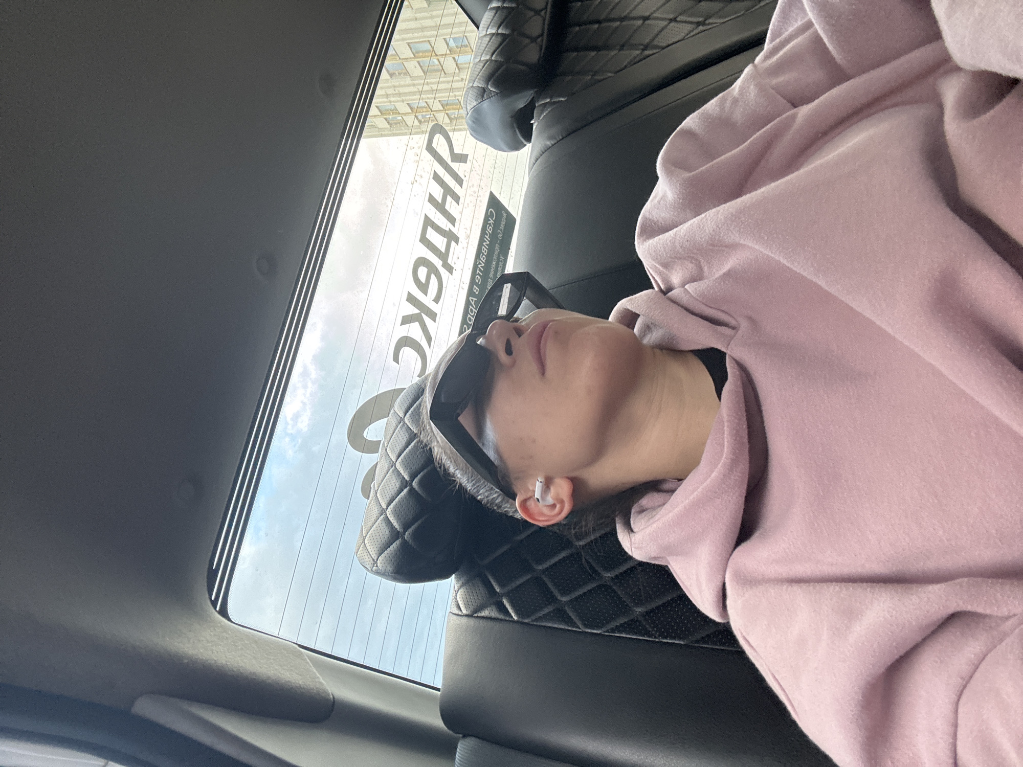 A person lies in the back seat of a car. The person is wearing sunglasses and a pink hooded sweatshirt. The back window shows a view of Moscow, Russia, including a sign that says "Такси" (Taxi). The person has a white earbud in their ear. The seat has quilted leather accents.