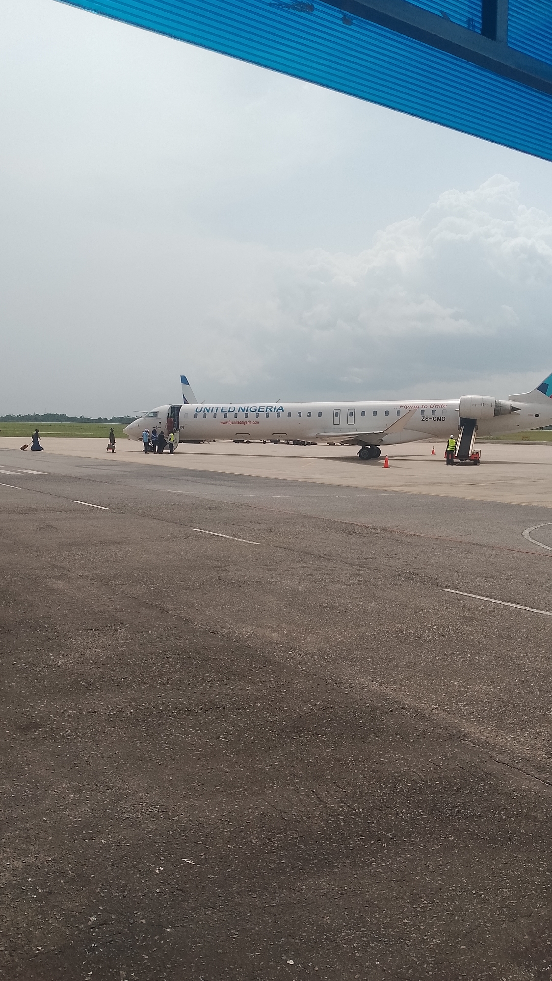 The image shows a United Nigeria aircraft, registration number ZS-CMO, on an airport tarmac. Passengers and ground crew are present near the aircraft. The location appears to be an airport, based on the presence of the plane, tarmac, and equipment. The city and region are Unknown. The sky is overcast.