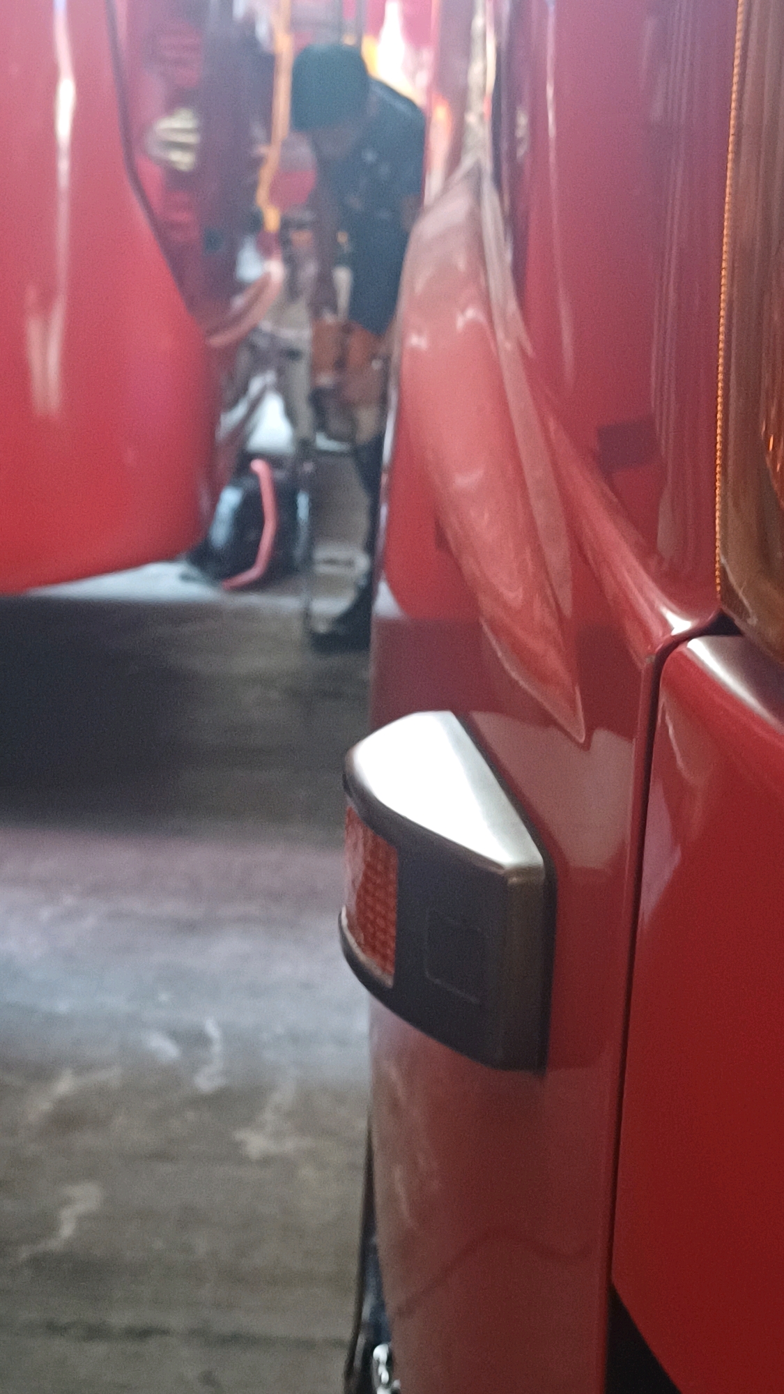 This is a close-up, low-angle shot of the rear corner of a red vehicle, possibly a van or truck, parked outdoors. The vehicle's bright red paint is highly reflective, creating a glossy appearance. A small, amber-colored reflector with a black housing is visible on the rear corner of the vehicle. In the background, a person wearing a dark t-shirt and a black baseball cap is leaning over, appearing to work on something within the vehicle or nearby. The ground appears to be concrete, with some debris or marks on its surface. The lighting suggests it is daytime, possibly in a workshop or garage setting. The exact location in South Jakarta, Indonesia, cannot be determined from the image alone.