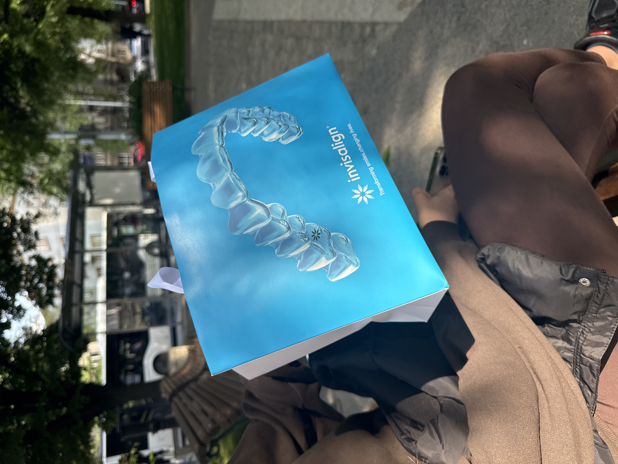 The image depicts a box for Invisalign aligners resting on a reflective surface, possibly a table. The box is predominantly blue with an image of clear aligners and the "Invisalign" logo. A portion of a person wearing brown leggings, a grey skirt, and a jacket is visible to the right of the box. In the blurred background, there are trees and what appears to be a street with buildings, potentially taken in an urban setting, but not verifiable to Plovdiv, Bulgaria.