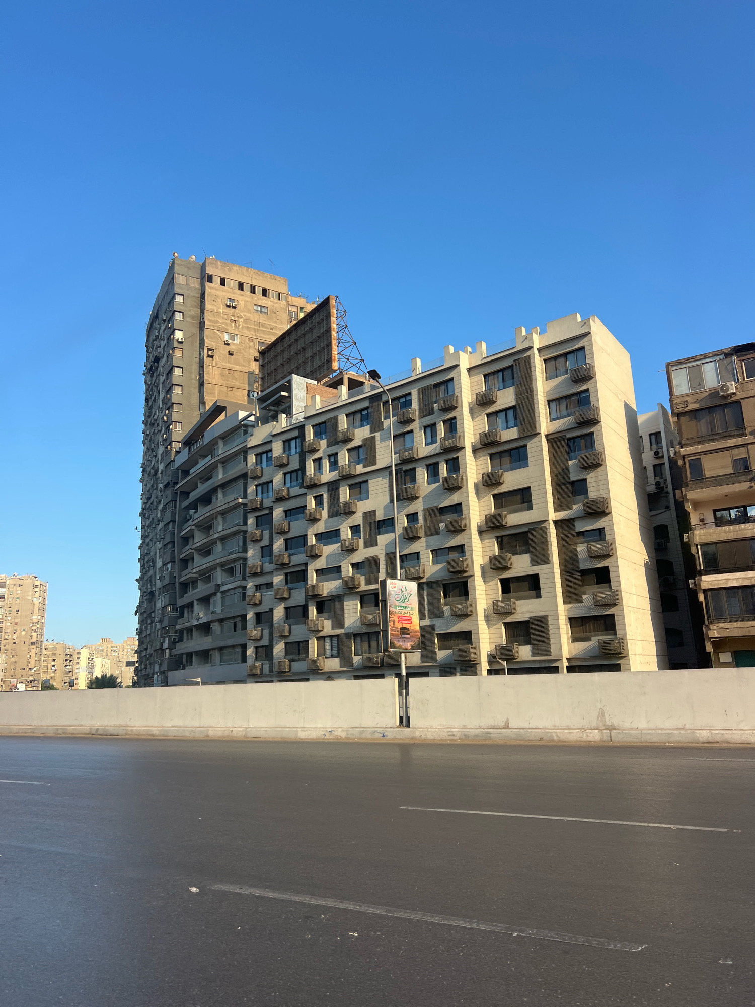 An exterior, low-angle shot captures a street view in Giza, Egypt, under a clear blue sky. The foreground features a paved road with a faint white lane marking. A low concrete barrier separates the road from an elevated walkway or bridge. Behind the barrier, a series of modern and older apartment buildings are visible. The most prominent building is a light-colored, multi-story apartment complex with numerous balconies, many equipped with air conditioning units. To its left, a taller, older, and darker building looms, showing signs of wear and featuring many windows and air conditioning units. Further to the left, more apartment buildings of varying architectural styles and ages are visible in the background. A large, faded billboard structure is also present atop one of the older buildings. A single lamppost and a street sign with Arabic text are positioned on the elevated walkway. The lighting suggests it is either early morning or late afternoon. No people are visible in the frame.