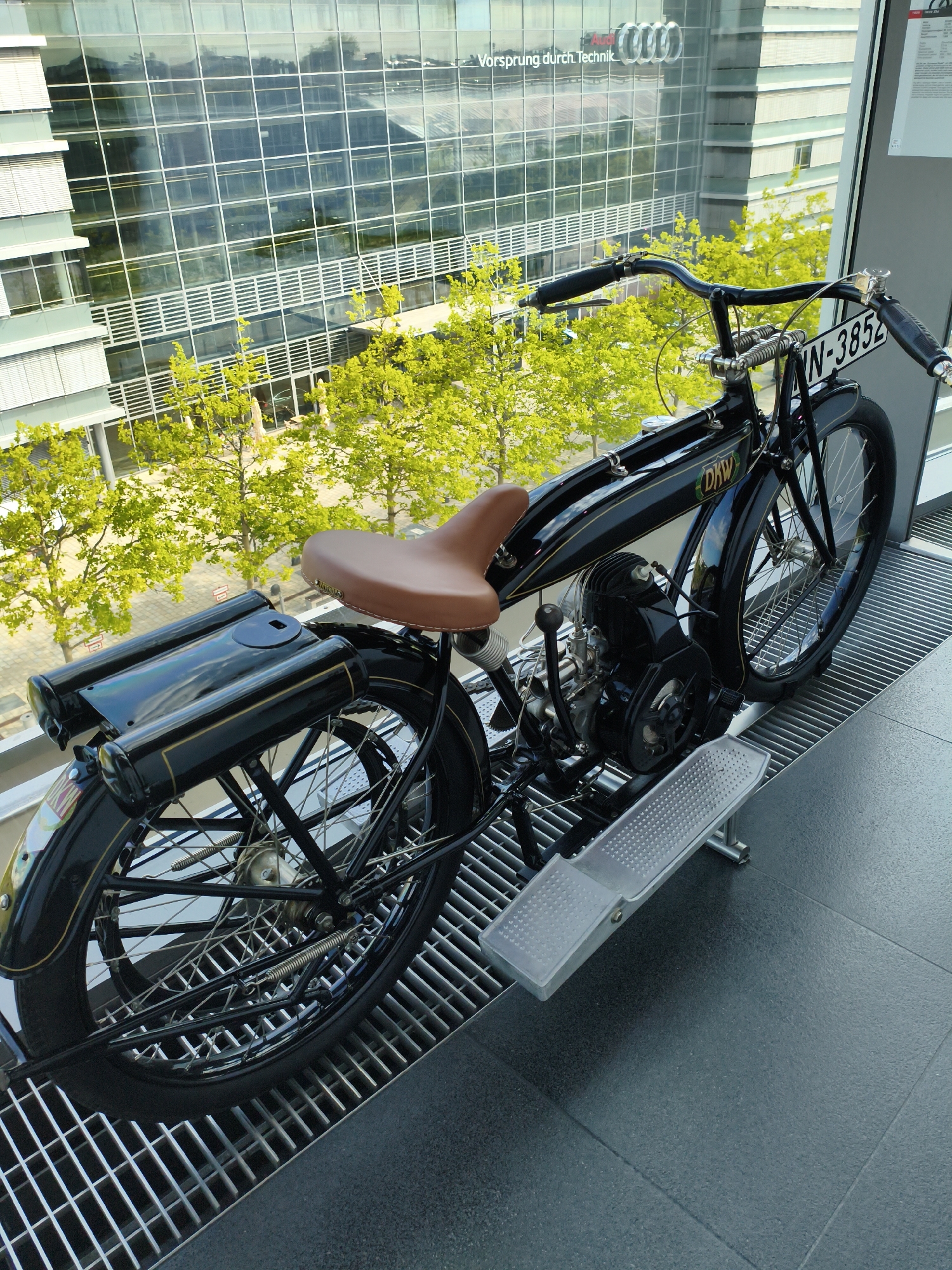 This image shows a vintage black DKW motorcycle displayed indoors, with a large glass-fronted building in the background. The building's facade features prominent Audi branding with the slogan "Vorsprung durch Technik." The motorcycle is black with a brown leather seat and has the license plate "IN-3852." It is positioned on a metal grate floor in front of a window that looks out onto trees and a street. The scene is likely within the Audi Forum in Ingolstadt, Germany, given the building's architecture and branding. There are no people visible in the image. The overall impression is of a historical exhibit showcasing automotive heritage.