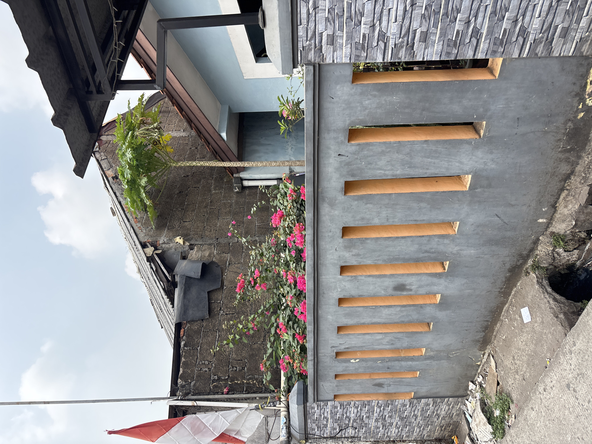 The image shows a residential scene in the city of Bekasi, Indonesia. A gray concrete fence with vertical orange-colored openings runs across the foreground. Behind the fence, a dense bush of bright pink bougainvillea flowers is in bloom. To the left of the flowers, a tall, thin papaya tree stands, and to its right, a light blue house with a dark corrugated metal roof is visible. A dark brick wall with some black sheeting attached is to the left of the papaya tree, and an Indonesian flag is mounted on a pole to the far left. The ground in front of the fence is paved, with debris and a damaged drain cover visible. The sky is overcast with white clouds.