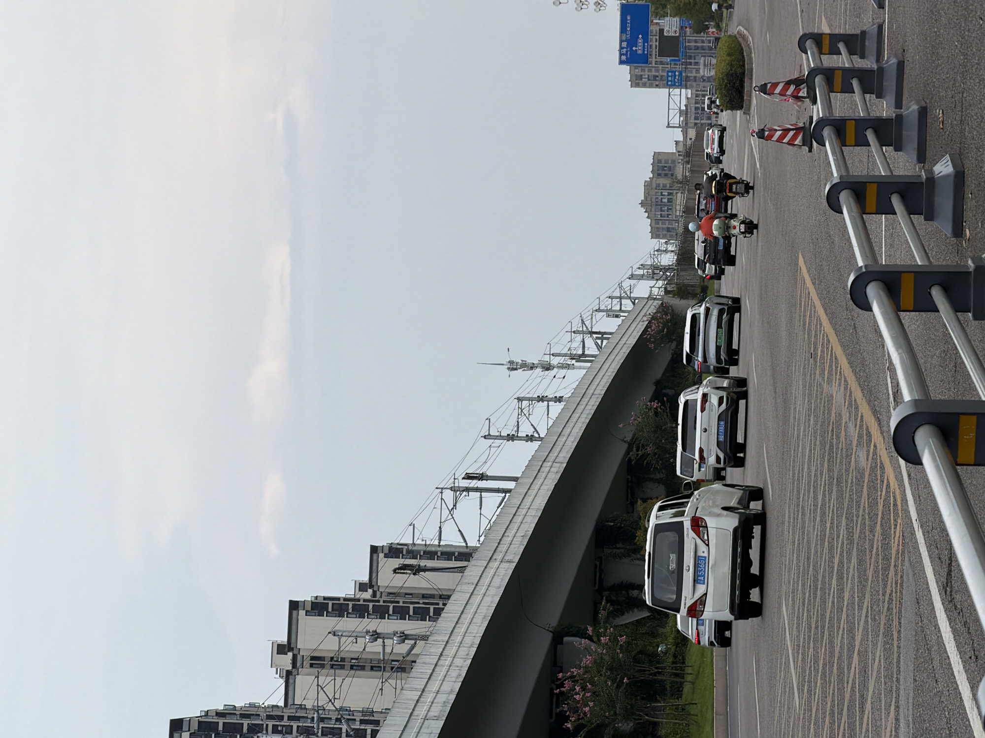 In Jiangjin, China, a multi-lane road runs beneath an elevated viaduct, likely for a train line, as evidenced by the overhead power lines and support structures. The road is populated with various vehicles including white cars, a white van with license plate "A 53561", and two motorcycles. One motorcycle, ridden by a person wearing an orange jacket, is in the foreground, with another motorcycle following behind. Traffic cones with red and white stripes are placed along the roadside barriers. In the background, modern apartment buildings and a taller, older-style building are visible against a cloudy sky. Road signs are present, indicating directions to various locations. The environment appears to be an urban setting during daylight.