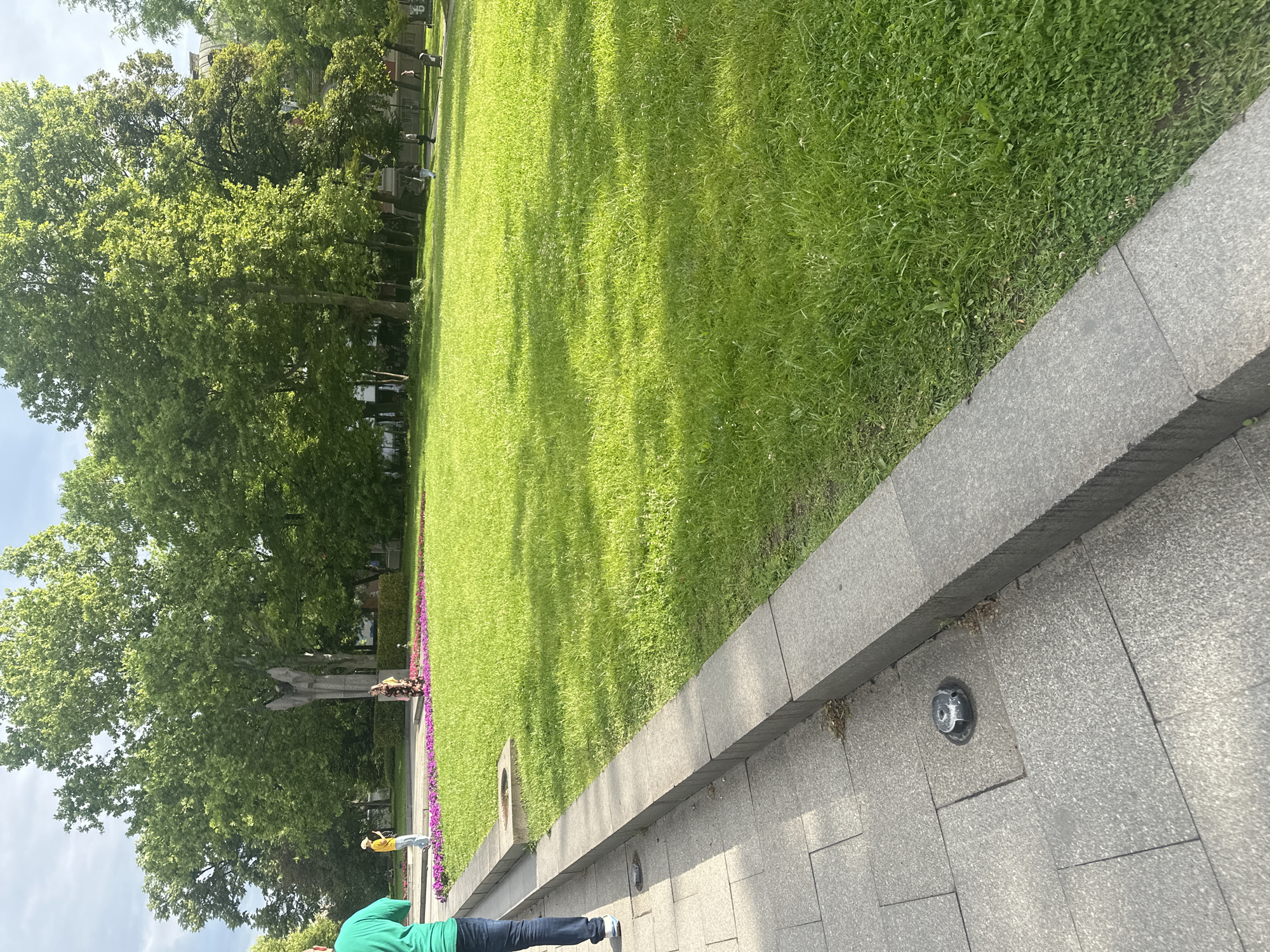 The image shows a park landscape in Sofia, Bulgaria, likely near the Monument to the Unknown Soldier. A large green lawn, partially shaded by trees, stretches across the scene. A granite wall with built-in lighting fixtures borders a paved walkway. A man wearing a green shirt and jeans stands on the walkway. In the distance, near the monument, another person wearing a yellow shirt stands. Purple flowers line the edge of the lawn near the monument.