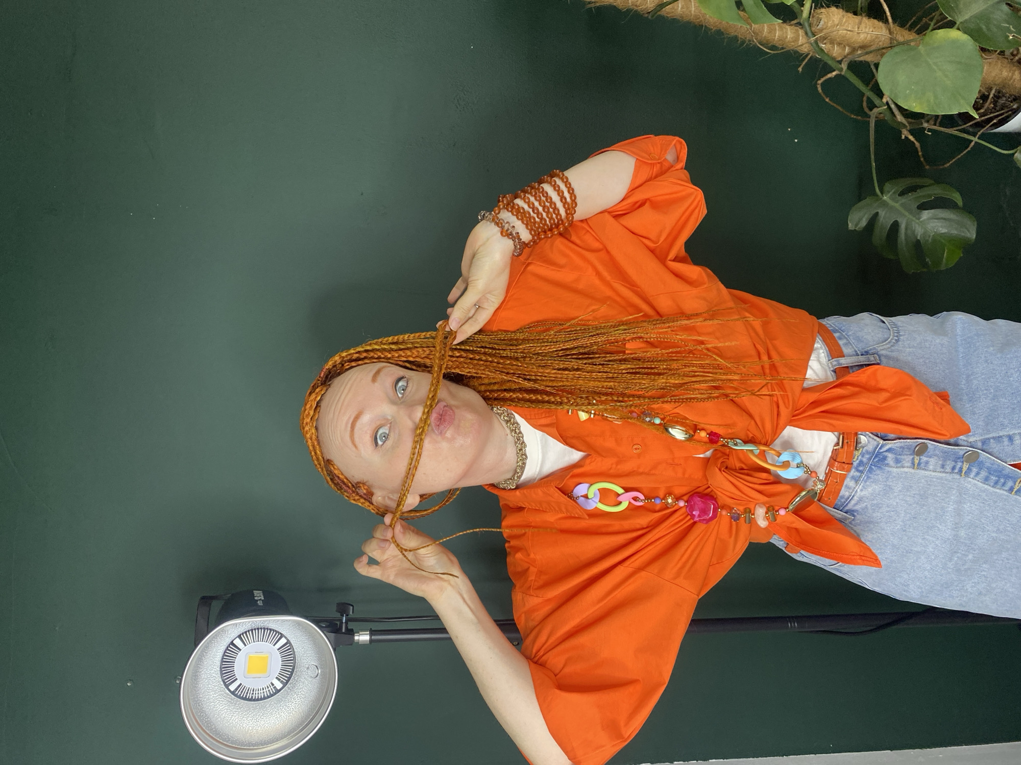 A woman with long, braided, reddish-orange hair and striking blue eyes is depicted in a playful indoor portrait. She is wearing an orange button-down shirt, tied at the waist, over a white undershirt, paired with light blue denim jeans and an orange belt. Her right arm is adorned with multiple strands of orange and brown beaded bracelets, and she wears a colorful, multi-piece necklace with various shapes and beads, as well as a gold-colored necklace underneath her shirt collar.
The woman is looking directly at the viewer with wide eyes and pursed lips, making an expressive, whimsical face. She is actively holding strands of her braided hair near her face with both hands.
The background consists of a solid dark green wall. To the upper right, a green plant with large leaves, possibly a Monstera, is visible, climbing on a coir pole. In the lower left, a studio light with a reflective dish and a central LED panel is positioned on a stand. Faint text "SLOW" is visible on the light's housing. The lighting suggests an indoor, controlled photography setup. The overall mood is vibrant and playful.