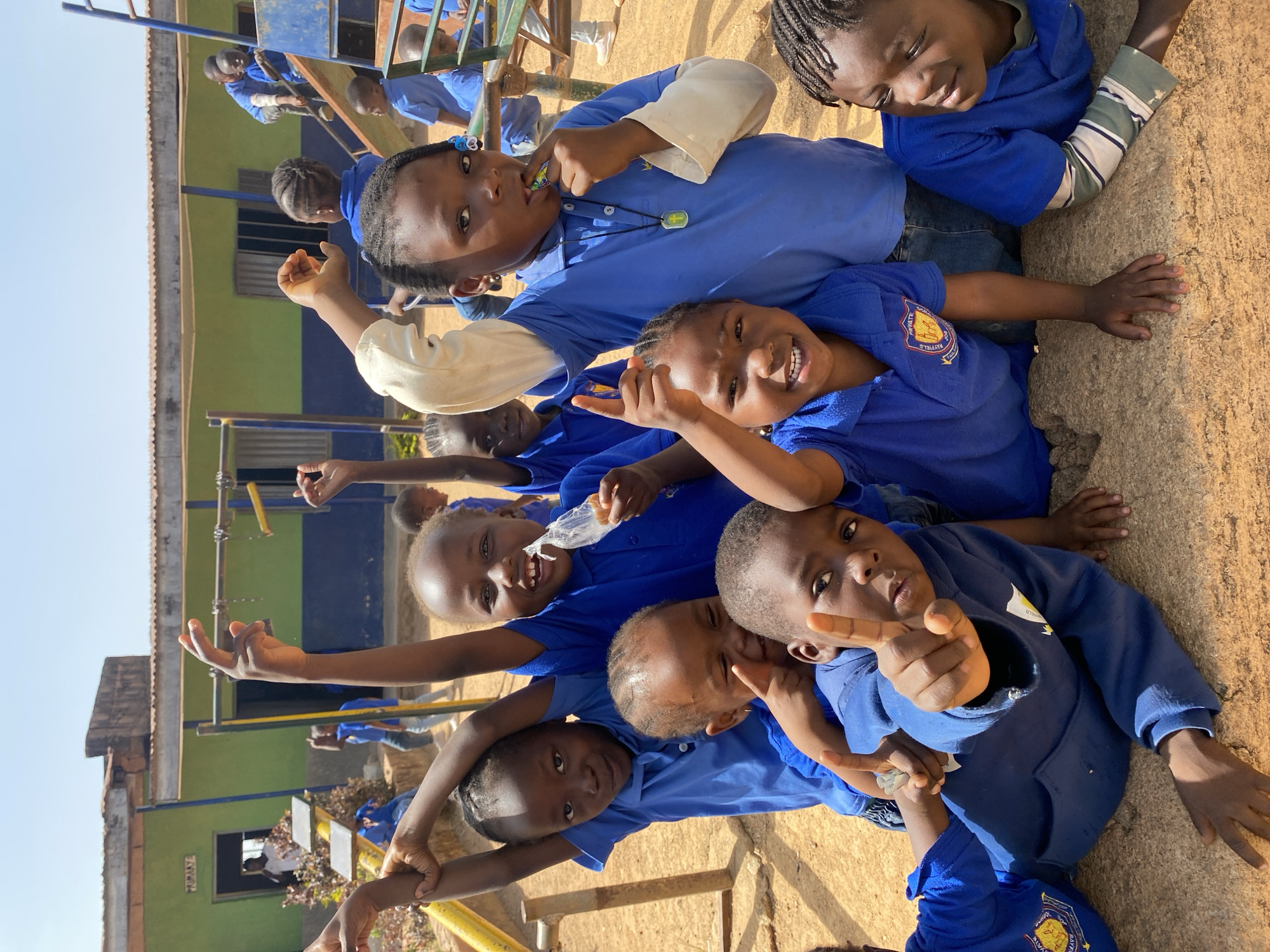 A group of school children, all wearing blue school uniforms, are seen outdoors in Gura Top, Nigeria. They appear to be on school grounds near playground equipment. They are smiling and interacting with each other, some are reaching their hands towards the camera. The background includes a building painted in green and blue. The scene is shot in daylight.