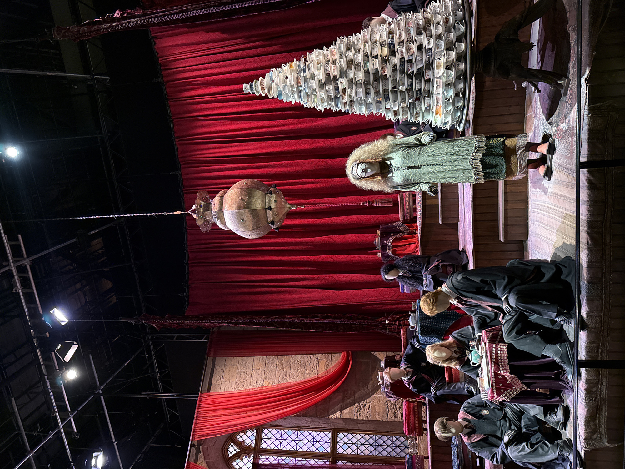 The image depicts a detailed set resembling a scene from the Harry Potter franchise, likely on display at Warner Bros. Studio Tour London – The Making of Harry Potter, located in Leavesden, Watford, United Kingdom.
The scene is set in an indoor studio environment, characterized by theatrical lighting from above and a dramatic backdrop of red velvet curtains. The flooring appears to be a combination of patterned rugs and raised wooden platforms, suggesting a staged display.
In the foreground, several mannequins dressed in Hogwarts robes are positioned around a small table set for tea. One mannequin is seated on the floor, leaning towards the table, while others are seated on cushions and benches. A teapot and teacups are arranged on the table, indicating a tea-drinking scene.
To the right of the mannequins, a surreal, towering structure made of stacked teacups and saucers dominates the space. A mannequin dressed in an elaborate, layered green costume with curly blonde hair and glasses stands beside the teacup tower.
In the background, more mannequins in robes are seated on raised platforms, and a large, ornate lantern hangs from the ceiling. A gothic-style window with stained glass is visible on the left side of the frame, further enhancing the magical atmosphere. The overall impression is a carefully curated exhibition designed to immerse visitors in the world of Harry Potter.