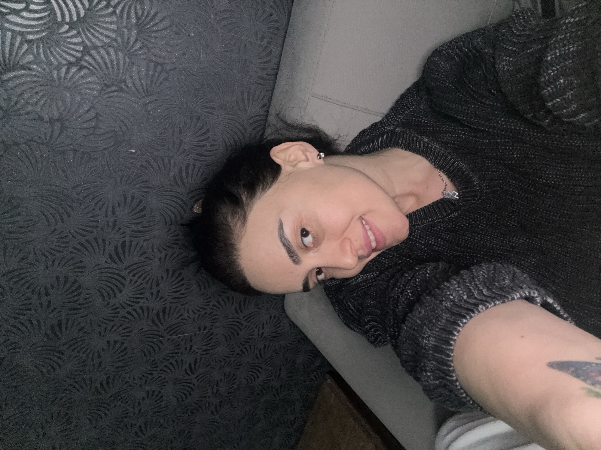 A person is reclining in an interior setting. The person has dark hair, fair skin, and prominent eyebrows. They are wearing a dark, knit sweater and a delicate necklace. A stud earring is visible in their left ear. A tattoo is partially visible on their left arm.
The person is reclining on a light gray upholstered surface, likely a sofa or lounge chair. To the left of the person is a textured gray surface with a repeating floral pattern. The image offers no information to establish the specific location in the city of Tbilisi, Georgia.