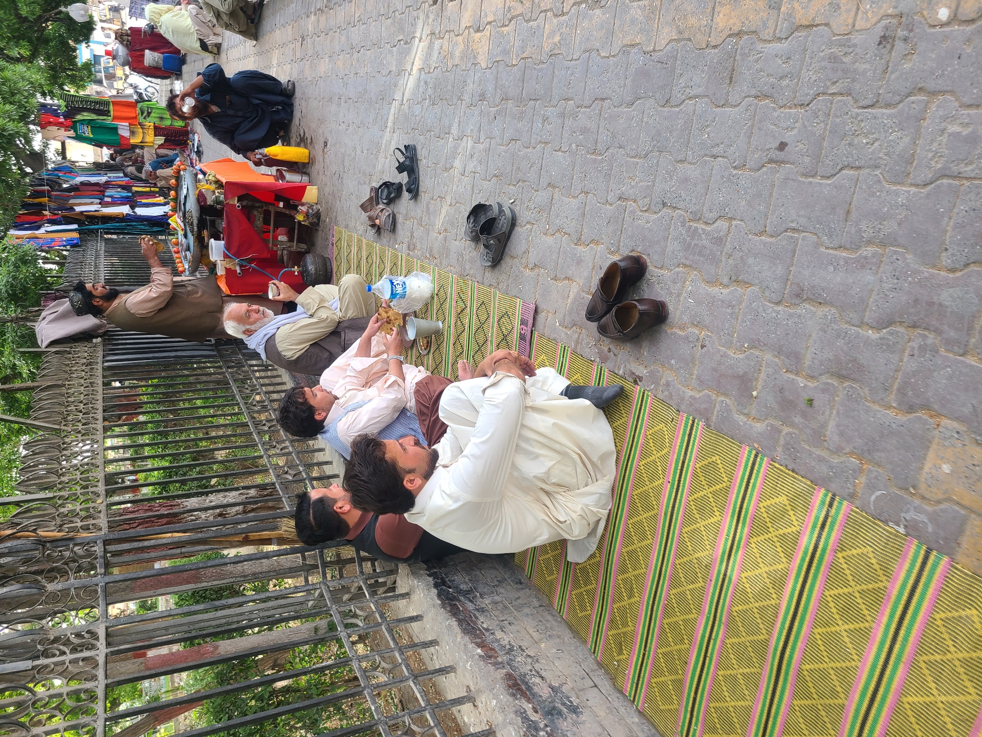 The image depicts a vibrant street scene, likely a market or a busy sidewalk, in Quetta City Tehsil, Pakistan. The perspective is tilted, showing the ground on the right and people and stalls on the left.
In the foreground, several men are resting on a long, patterned mat featuring yellow, green, and pink stripes, laid out on grey interlocking paving stones. They are dressed in traditional South Asian attire, primarily shalwar kameez. Some men are lying down, while others are seated, appearing relaxed. One man is holding what looks like a piece of bread or paratha and a plastic cup, while another plastic water bottle, labeled "WATER," rests nearby. A pair of dark shoes is placed neatly beside the mat, and other shoes and sandals are scattered further along the paved area.
To the left, a black metal fence separates the resting area from what appears to be a green, treed space, possibly a park or garden. Beyond the fence and the resting men, the street becomes a marketplace. Several vendor stalls are visible, piled high with colorful clothing and other goods. One vendor is operating a cart that has oranges displayed on its side and various items for sale. Another man in dark blue clothing is squatting near this cart, seemingly interacting with the vendor. Further into the background, more people and stalls are visible, indicating a bustling environment.
The scene takes place during daylight hours, under what appears to be an overcast or diffused sky, as there are no harsh shadows. The overall impression is one of everyday life in a public urban setting, where people gather, trade, and rest.