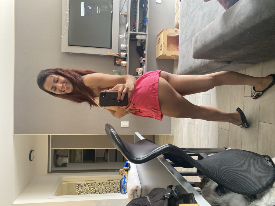 A woman stands in front of a mirror, taking a selfie. She is wearing a pink skirt and flip-flops. The background features a living room with a TV, shelves, and a couch. A black chair is positioned in front of the mirror, partially obscuring the view.