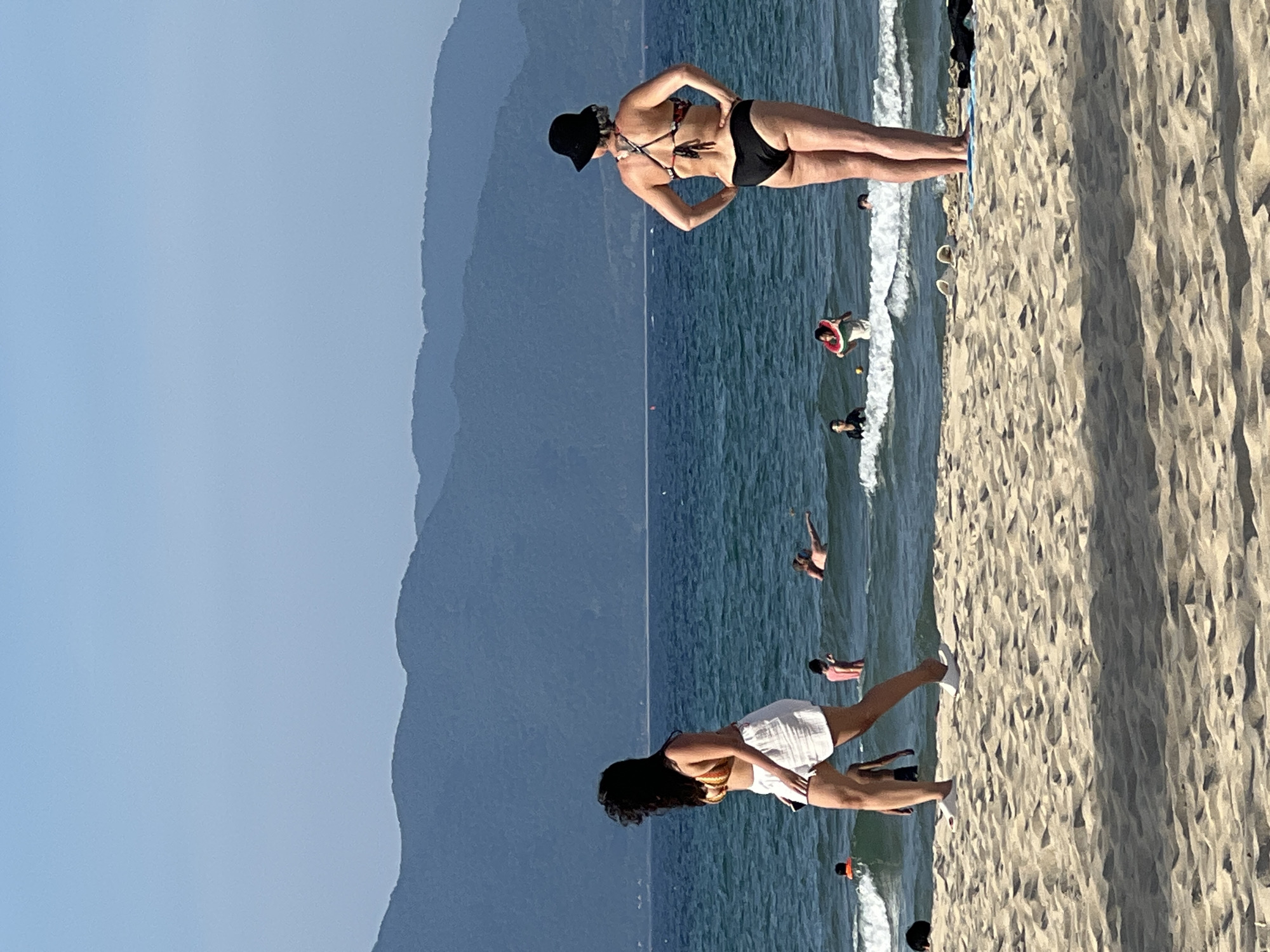 The image shows a beach scene with several people in the ocean and on the shore, with a mountain range in the background. The scene is likely located in Da Nang, Vietnam, given the prevalence of similar beaches and the mountainous terrain.
In the foreground, the sandy beach is visible with footprints and shadows. Two women are prominent: one in the left mid-ground walking towards the left, wearing a striped bikini top and white shorts. Her hair is dark and wavy. Another woman is in the right mid-ground, facing away from the camera, wearing a black bikini and a black hat. Her hands are on her hips.
In the ocean, several people are visible. Some are wading, while others are swimming or floating. One person is wearing an orange floatation device, and another is holding a watermelon-shaped inner tube. The water is a clear blue, with small waves breaking near the shore.
The background features a range of blue-hued mountains, indicative of a coastal landscape. The sky is clear and bright blue, suggesting a sunny day. The overall impression is of a relaxed, warm weather day at a popular beach destination.