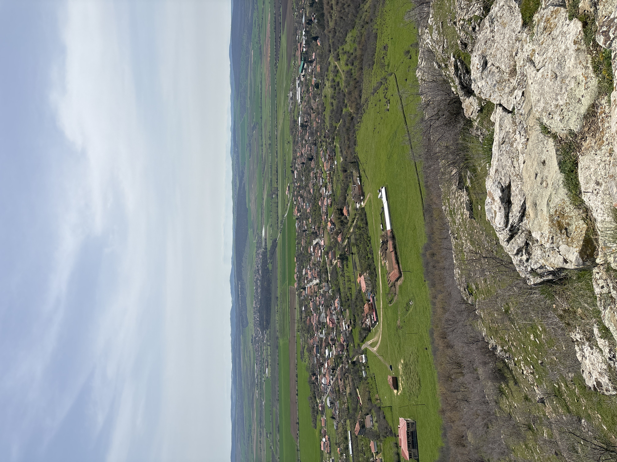 The image is a landscape view of a rural area near Goritsa, Bulgaria. The camera is positioned high, possibly from a clifftop or elevated viewpoint. In the foreground, there are large, greyish-white rocks and dry vegetation, indicative of a rocky terrain.
A wide, green valley stretches out from the foreground, containing a village nestled within it. The village consists of numerous houses with red-tiled roofs, scattered along a road that runs through the valley. The valley is bordered by green fields, some of which are cultivated, and patches of trees. In the background, there is a distant view of hazy hills and sky, indicating the horizon line. The sky is overcast with grey clouds.