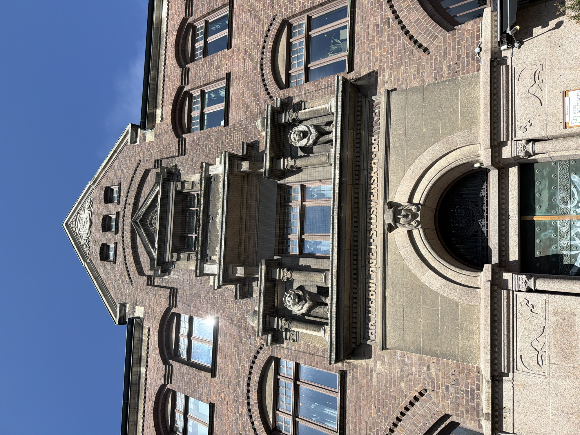 The image shows the facade of a brick building in Lund, Sweden. The building features arched windows and decorative lion sculptures on the upper levels. Below these, the words "AKTIEBOLAGET SYD SVENSKA BANKEN" are inscribed. The main entrance below is an arched doorway with ornate metalwork. The sky is clear and blue.