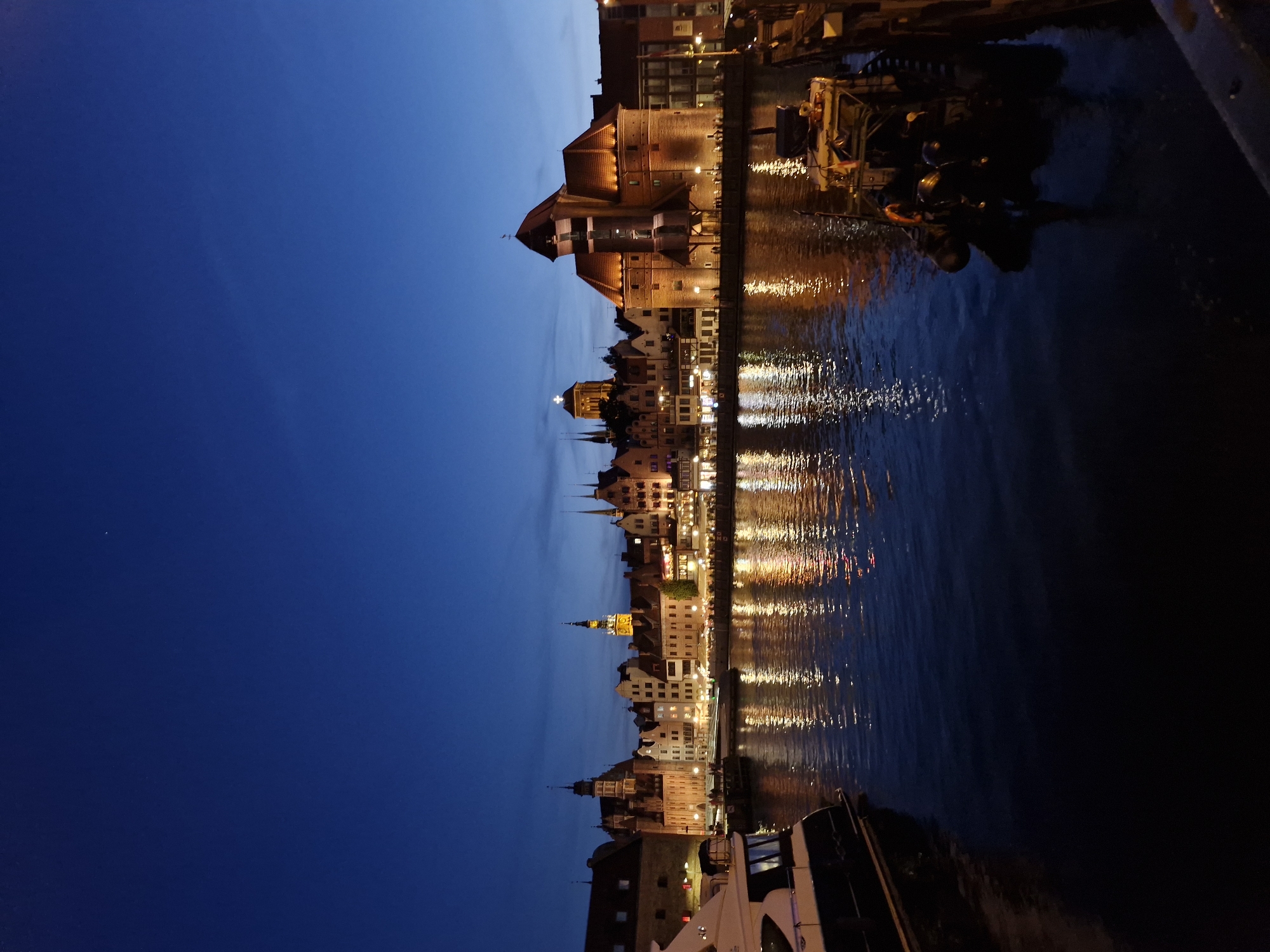A twilight view of the waterfront in Gdansk, Poland showcases the city's historic architecture, illuminated by warm artificial lights. The sky is a deep blue, transitioning into a lighter hue towards the horizon. A tranquil body of water reflects the city lights, creating a shimmering effect on the surface. In the foreground, a modern white yacht is docked on the left, its sleek design contrasting with the traditional buildings. To the right, a smaller boat with visible equipment and people is moored. The architecture features a variety of buildings, including a prominent brick structure with a red-tiled roof and several taller buildings with spires, characteristic of European cityscapes. The scene suggests a peaceful evening in a city steeped in history.