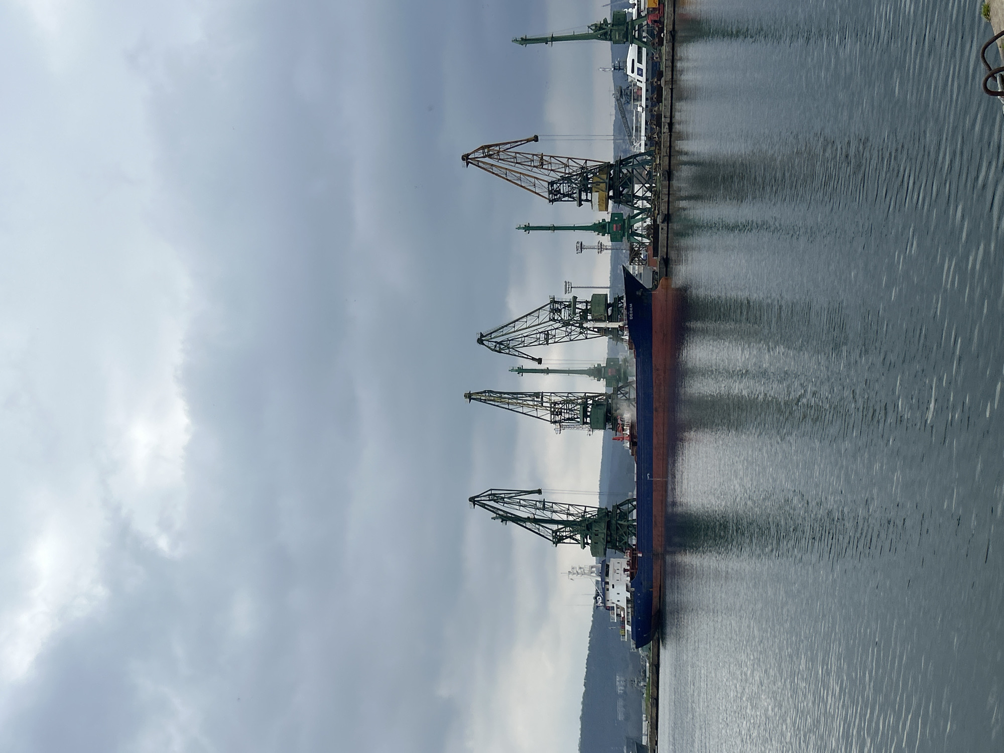 The image depicts a waterfront scene with multiple cranes along a harbor or dock. A large vessel is docked alongside the quay, seemingly in the process of being loaded or unloaded. The water is calm with reflections. The sky is overcast. The location is likely in the city of Unknown, Unknown. The scene is industrial.