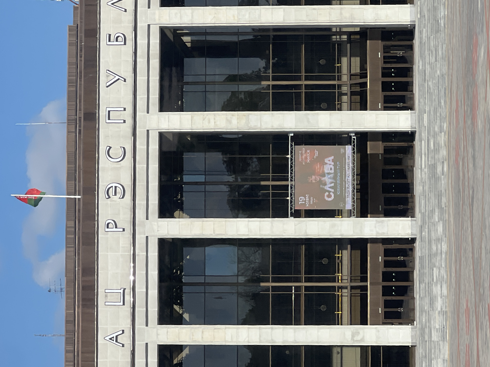 The image shows the exterior of the Palace of the Republic in Minsk, Belarus, on a sunny day. The facade is made of stone with large glass windows and multiple entrances. Above the entrances, the words "РЭСПУБЛІКА" (Republic) are inscribed in large, white, block letters. A Belarusian flag flies from a flagpole on the roof. A banner hangs in one of the central windows, advertising an event. The ground in front of the building is paved with stone tiles and there are wide stone steps leading up to the entrances. The sky is blue with some white clouds.