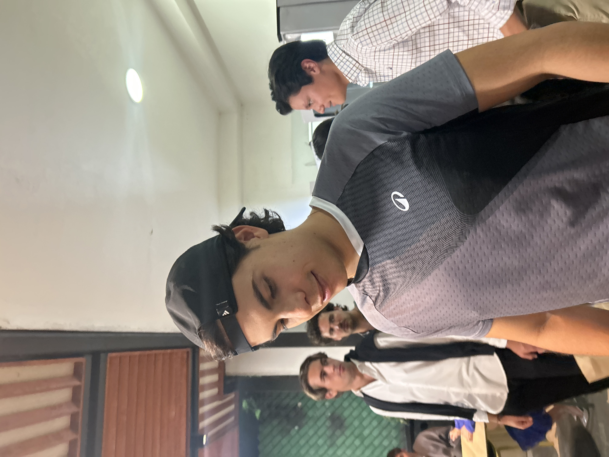 The image shows a group of people in an indoor setting. A person wearing a black Adidas baseball cap and a gray and black polo shirt is in the foreground, with others partially visible behind. One person behind is wearing a white shirt with a black stripe down the front. Another person is wearing a white and brown checkered shirt. The location is identified as Unknown, Unknown. The background shows a wall with a small, round window-like light fixture. Part of a wooden structure is also visible in the background.