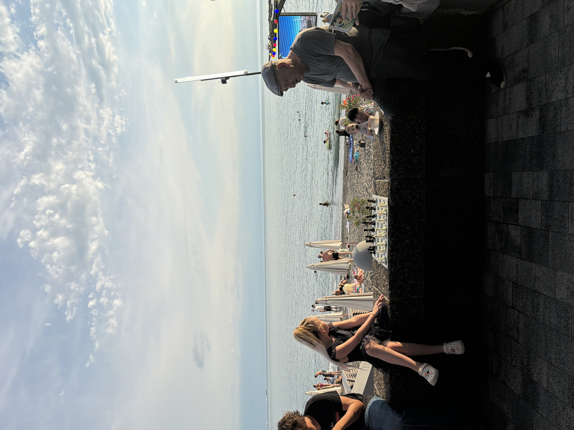 This is a medium shot of a coastal scene in Sochi, Russia, on a bright day with scattered clouds. In the foreground, on the left, a young man with dark curly hair wearing a black t-shirt and jeans sits facing left, his arm resting on his knee. Next to him, a young woman with blonde braided hair, wearing a black frilly dress and white clogs, sits facing right, her hands poised over a chessboard. The chessboard is set up on a low, dark stone wall.
Behind them, the beach is populated with people sunbathing, swimming, and walking. Beach umbrellas are scattered across the sand. The sea stretches out to the horizon, with a few boats visible in the distance. On the right side of the frame, an older man wearing a grey t-shirt and a flat cap sits facing left, looking towards the sea. He is holding a book or a tablet. Partially visible to his right is a person in a light-colored jacket, holding a white bag. A large, illuminated screen displaying a vibrant image is visible in the background on the right, suggesting an entertainment venue. A tall, thin light fixture stands near the screen. The overall atmosphere is relaxed and summery.