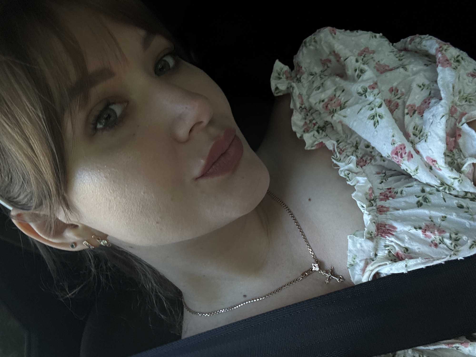 A close-up selfie of a young woman with light brown hair and fair skin. She is looking towards the camera with a slight smile. Her eyes are green and her lips are painted with a muted pink lipstick. She is wearing a white off-the-shoulder top with a floral pattern of pink roses and green leaves. A delicate rose gold necklace with a cross pendant is visible on her neck. Her hair is pulled back slightly, revealing small stud earrings in her left ear. A black seatbelt is visible across her chest, suggesting she is in a vehicle. The background is dark, with some strands of her hair gently blowing across her face. There is no indication of the specific location within Chaikovsky, Russia, in the image.