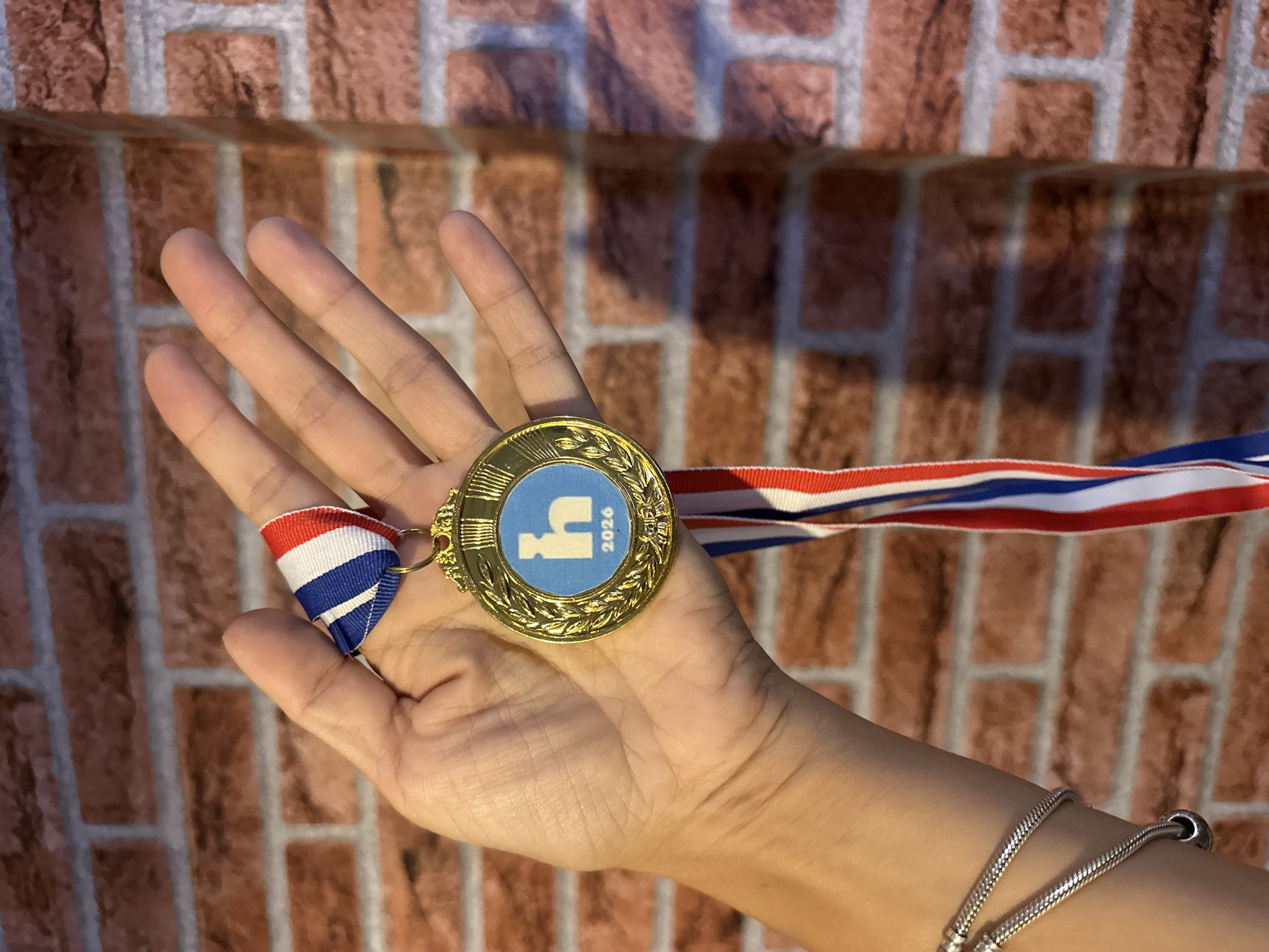 A close-up, eye-level shot shows a person's open right hand holding a gold medal on a red, white, and blue ribbon. The medal has a laurel wreath design around the edge and a blue circle in the center with a white stylized "h" and the number "2026" below it. The hand is positioned in front of a brick wall with a mottled reddish-brown and grey pattern. The background is blurred, suggesting the image was taken in an indoor environment with artificial lighting. The scene does not provide information about its specific location in Kathu, Thailand.