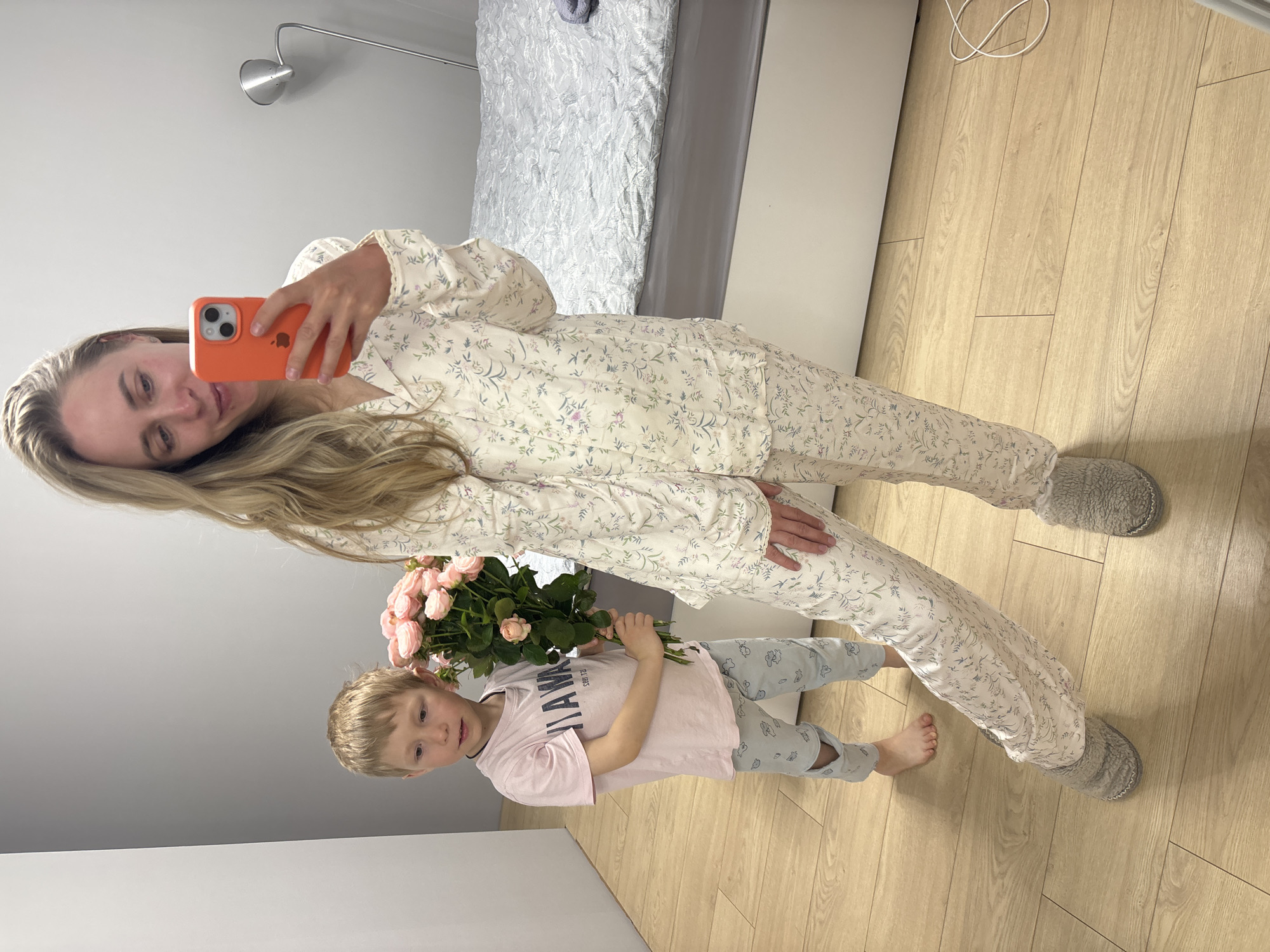 A woman is taking a selfie with a young child in what appears to be an indoor setting. The woman is wearing floral pajamas and gray slippers, and she is holding an orange smartphone. The child, who is also in pajamas, is holding a bouquet of pink roses. The room features wooden flooring and a bed in the background with light bedding. The overall scene suggests a domestic setting, possibly a bedroom. There is no explicit information to confirm that the location is in Chaikovsky, Russia, or anywhere else for that matter.