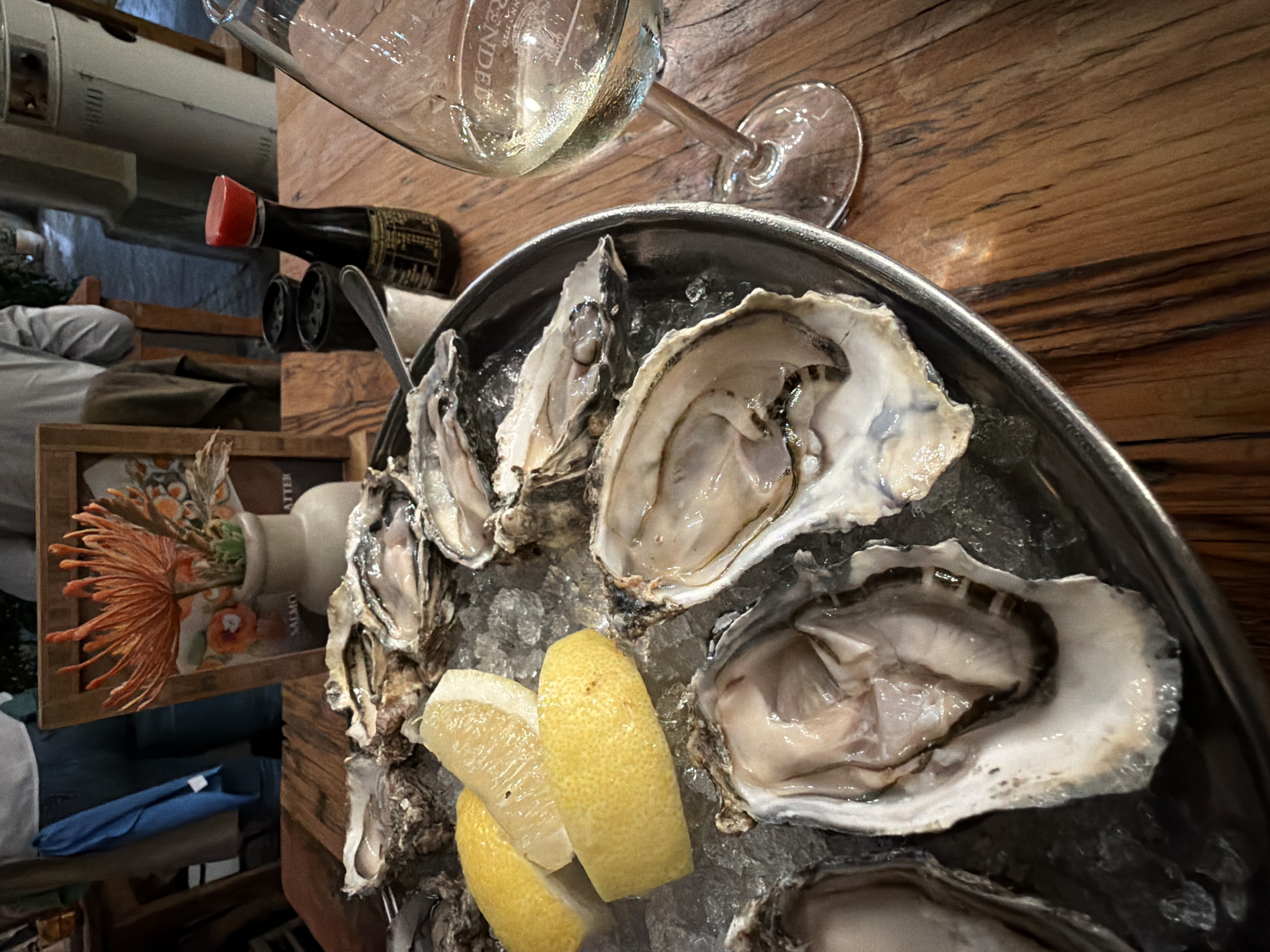 The image depicts a platter of oysters on ice with lemon wedges, accompanied by a glass of white wine and a small bottle of sauce, likely soy sauce or a similar condiment. The scene is set on a wooden table, with a vase of flowers and other restaurant-like items visible in the background. Considering the common culinary pairing of oysters and wine, this scene likely takes place at a restaurant or bar.
While precise forensic identification of the location as Cape Town, South Africa cannot be definitively confirmed solely from visual elements in the image, the setting and the presence of oysters are indicative of a coastal city with seafood offerings. Without other definitive contextual clues, however, absolute confirmation of the location as Cape Town isn't possible.