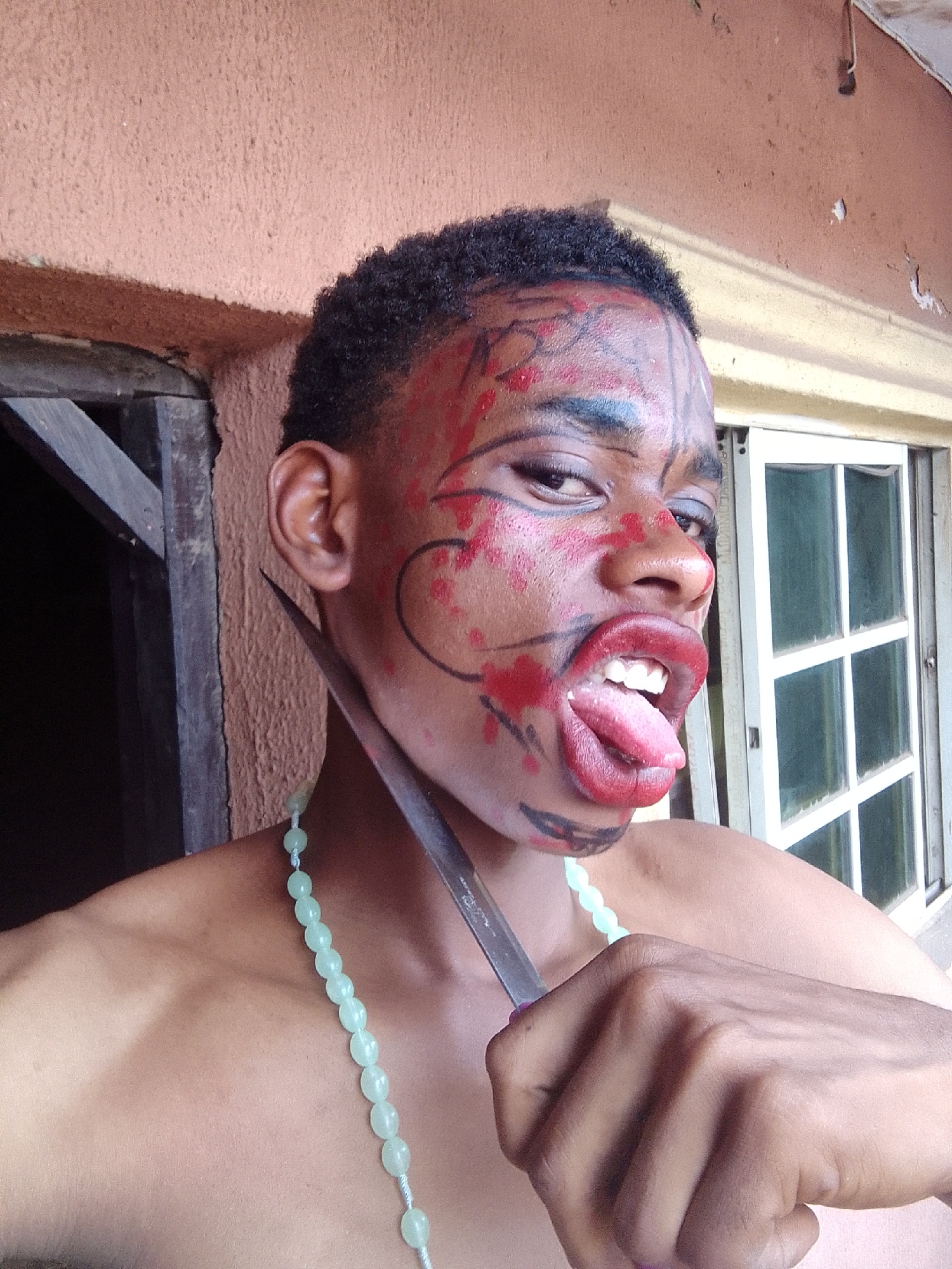 A young individual, appearing male with dark skin and short, curly black hair, is captured in a dramatic, close-up portrait. The person is bare-chested and wears a light green beaded necklace. Their face is covered in theatrical makeup, featuring black lines and red splotches that resemble tribal markings or blood, especially around the forehead, cheeks, and nose. Red lipstick is applied messily around the mouth, which is open wide with the tongue fully extended, exposing teeth. The individual's right eye, marked with dark eyeliner, gazes directly at the viewer, while the left eye is partially obscured by the angle and makeup.
A metallic, sharp-bladed knife is held in the person's right hand, with the blade resting against their neck, just below the left ear. The pose, makeup, and expression collectively convey an intense, wild, or possibly menacing character.
The setting appears to be outdoors or in an open doorway, with a textured, light reddish-brown wall forming the background. To the left, dark wooden beams or framing are visible. To the right, a white-framed window with multiple panes is present. The lighting is bright and natural, suggesting it is daytime. No discernible text is visible in the image.
