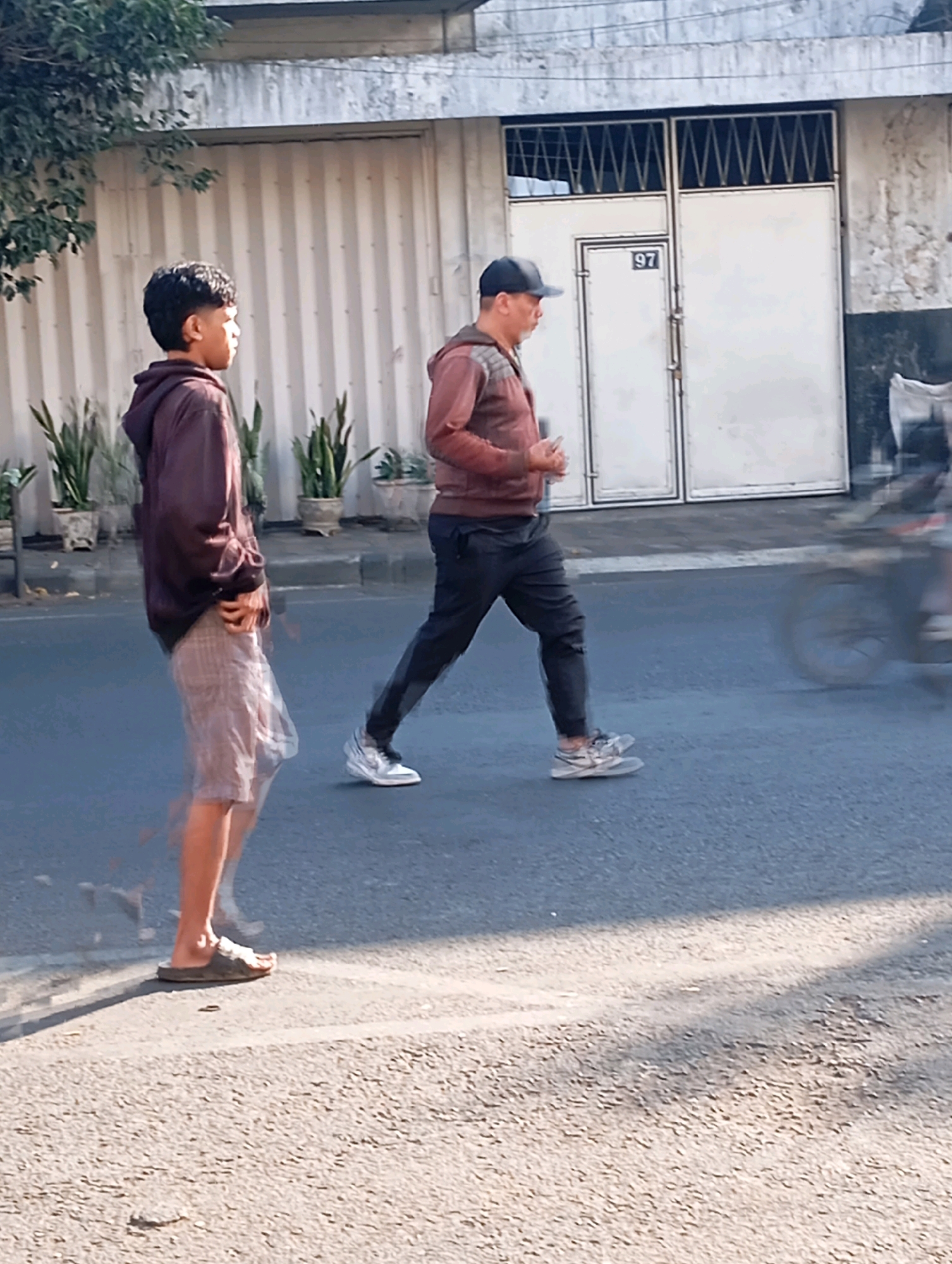 The image shows two males walking on a street in front of a building with a corrugated metal wall and double white doors with the number "97" above the left door. The building appears to be in a city. The male on the left, who is younger, is wearing a maroon hoodie and plaid shorts and sandals. He is captured with motion blur, suggesting movement. The male on the right, who is older, is wearing a maroon hooded jacket, dark pants, and athletic shoes. He is also in motion. A motorcycle is visible in motion in the background on the right side of the image, also showing motion blur. The ground is paved asphalt. The lighting suggests it is daytime. The presence of a building with a numbered door and paved street indicates an urban or semi-urban environment. Without further contextual information, pinpointing the exact location within Malang, Indonesia is not possible, but the scene depicts a common street view.