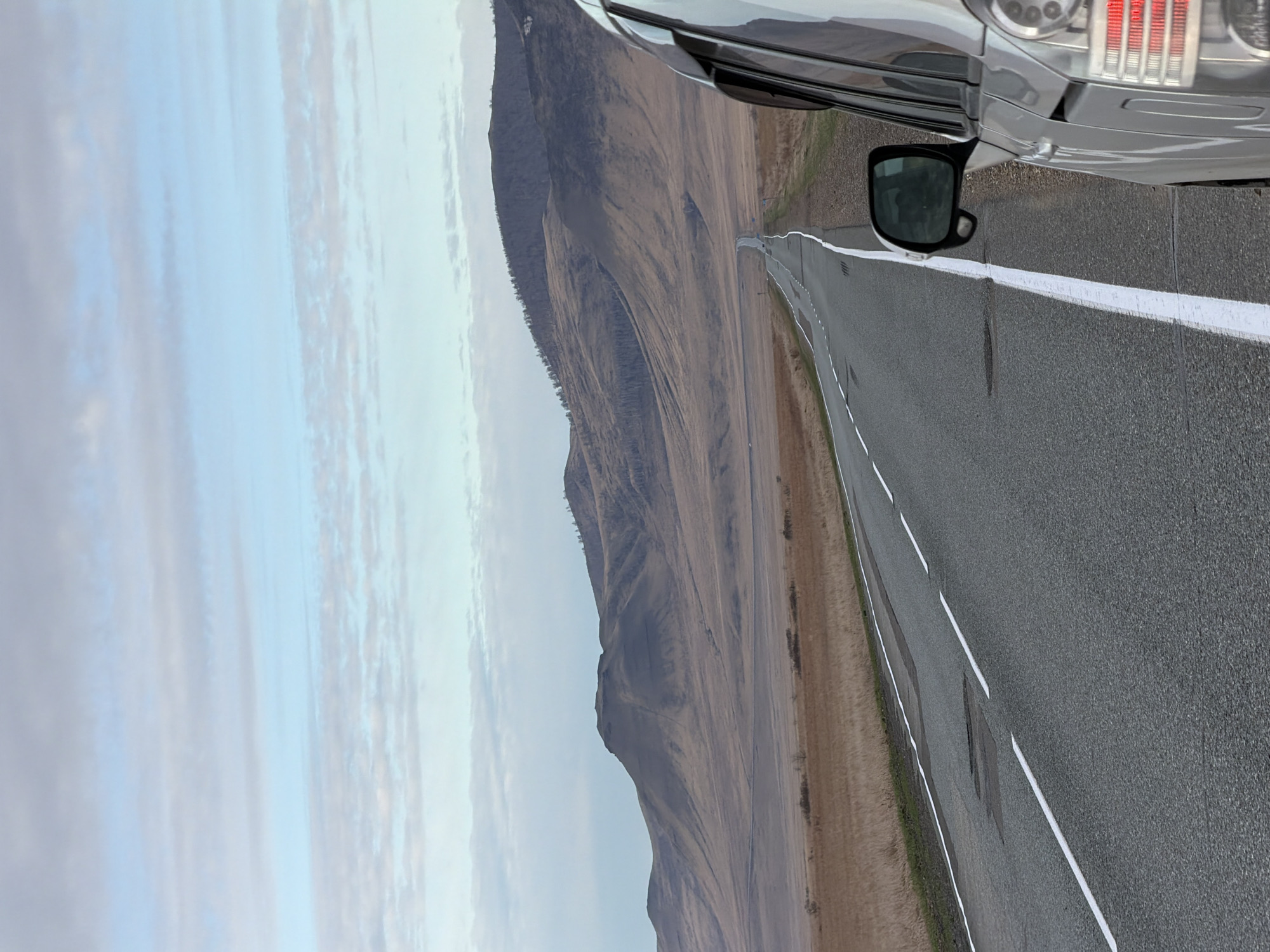 The image shows a gray car parked on the shoulder of a two-lane asphalt road. A white line marks the road's edge. The landscape features a large, sloping hill or mountain covered in dry, brown vegetation. The sky is overcast with light blue and gray clouds. The location is rural, consistent with an area outside a city, and unlikely to be within Aeshka, Russia. No people are visible.