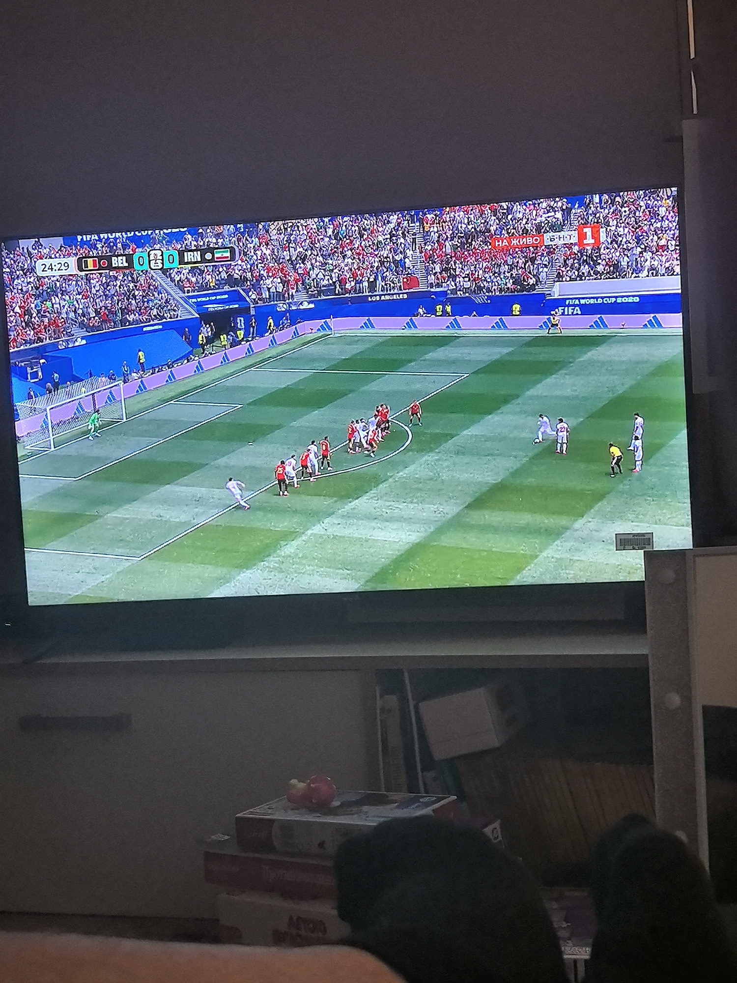 A television screen displays a live soccer match. The score is 0-0 between BEL and IRN, with the game at 24:29. The stadium is packed with spectators wearing red and white. A banner in the stadium reads "НА ЖИВО - БЪЛГАРСКИ" and displays the number 1. The soccer field is green with white lines. Players in red and white uniforms are visible on the field, with one player in a blue uniform. The scene is shot from a low angle, implying the camera is near the floor. In the foreground, part of a leg is visible, suggesting someone is watching the television. The bottom of the screen shows a shelf with stacked boxes, and some household items are scattered on the shelf.