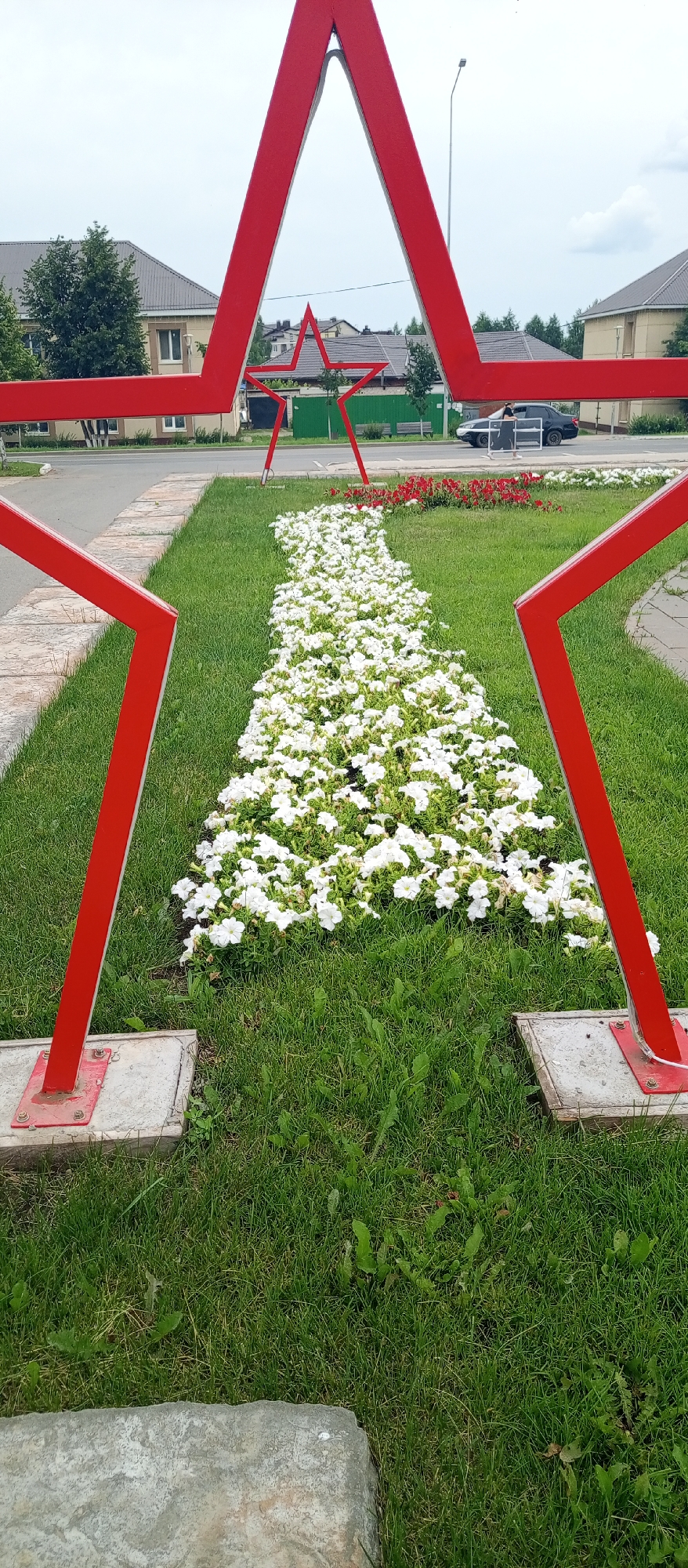 In Agryz, Russia, a park features a large, red, five-pointed star sculpture. The sculpture's base rests on concrete slabs on a grassy area. In front of the sculpture, a flower bed is densely planted with white flowers, with a smaller cluster of red flowers visible further back. Beyond the park area, there's a road with a black car driving and a person standing near it. Residential buildings with neutral-colored walls and dark roofs are visible on the left and right sides of the scene, separated by trees and green fences. The sky is overcast.