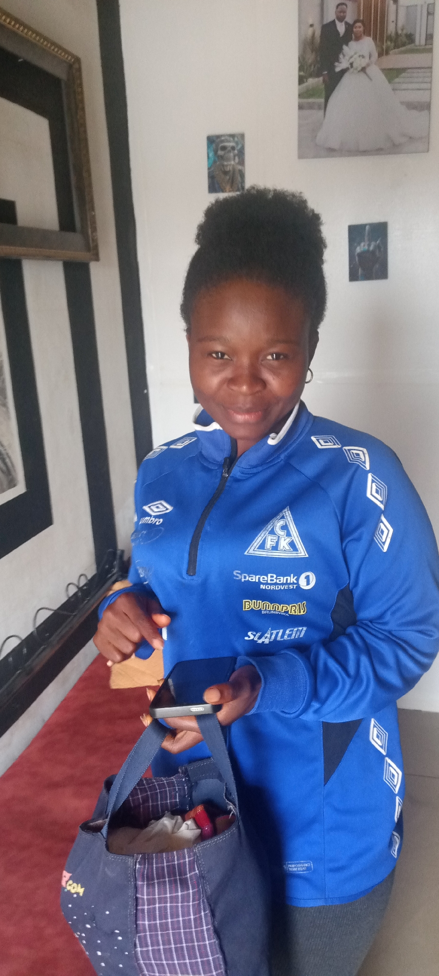 A medium shot depicts a woman holding a mobile phone in one hand and what appears to be a small wooden object in the other, along with a tote bag. The woman is wearing a blue athletic jacket with logos and text on the front. Behind her, the wall is decorated with framed photos. The scene is taking place indoors; the location and city cannot be determined from the image provided.
