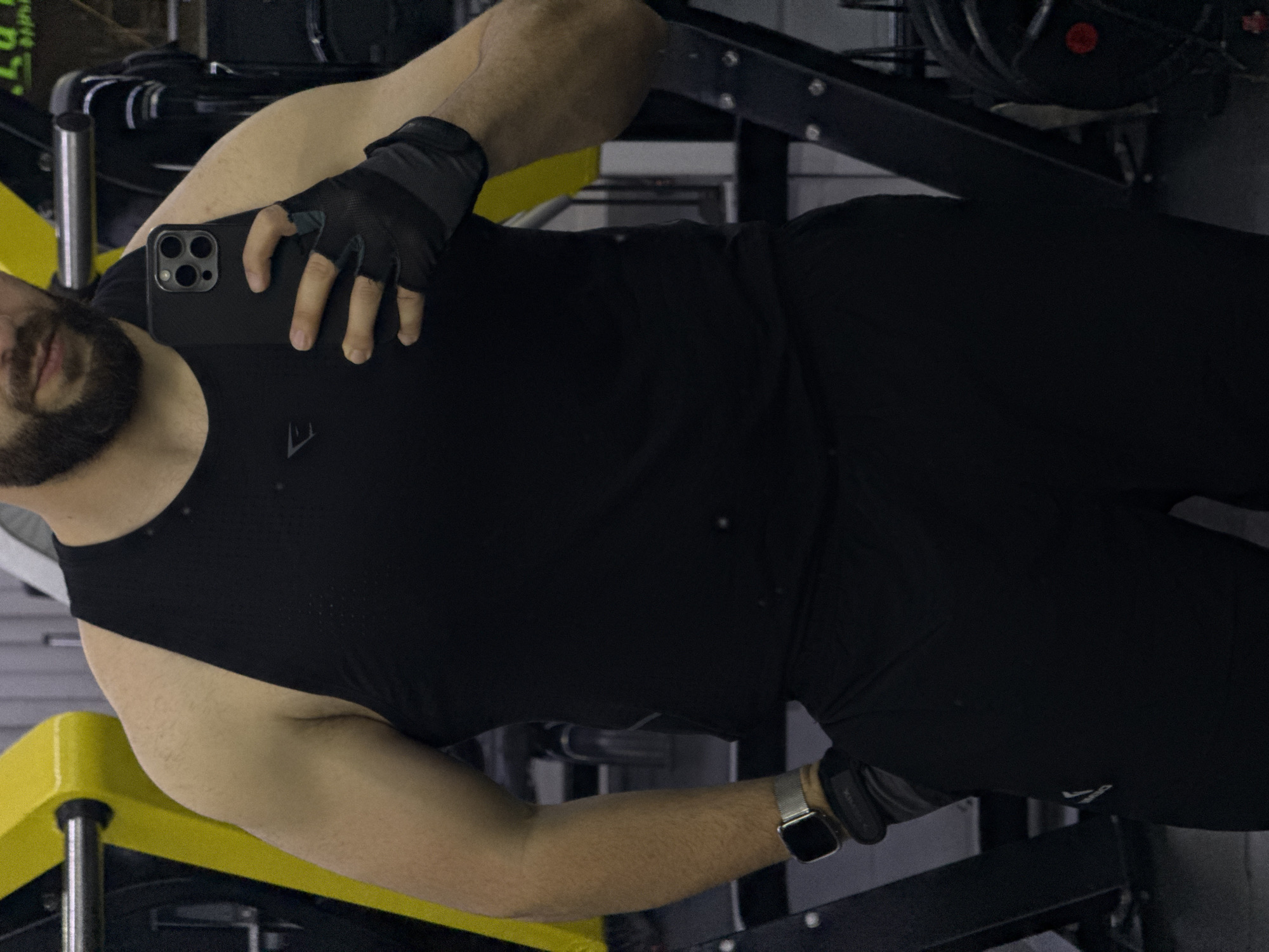 A person with dark hair and a beard is taking a selfie in a gym. They are wearing a black tank top, black gloves, and a watch. The gym has yellow and black equipment. The location is Yerevan, Armenia.